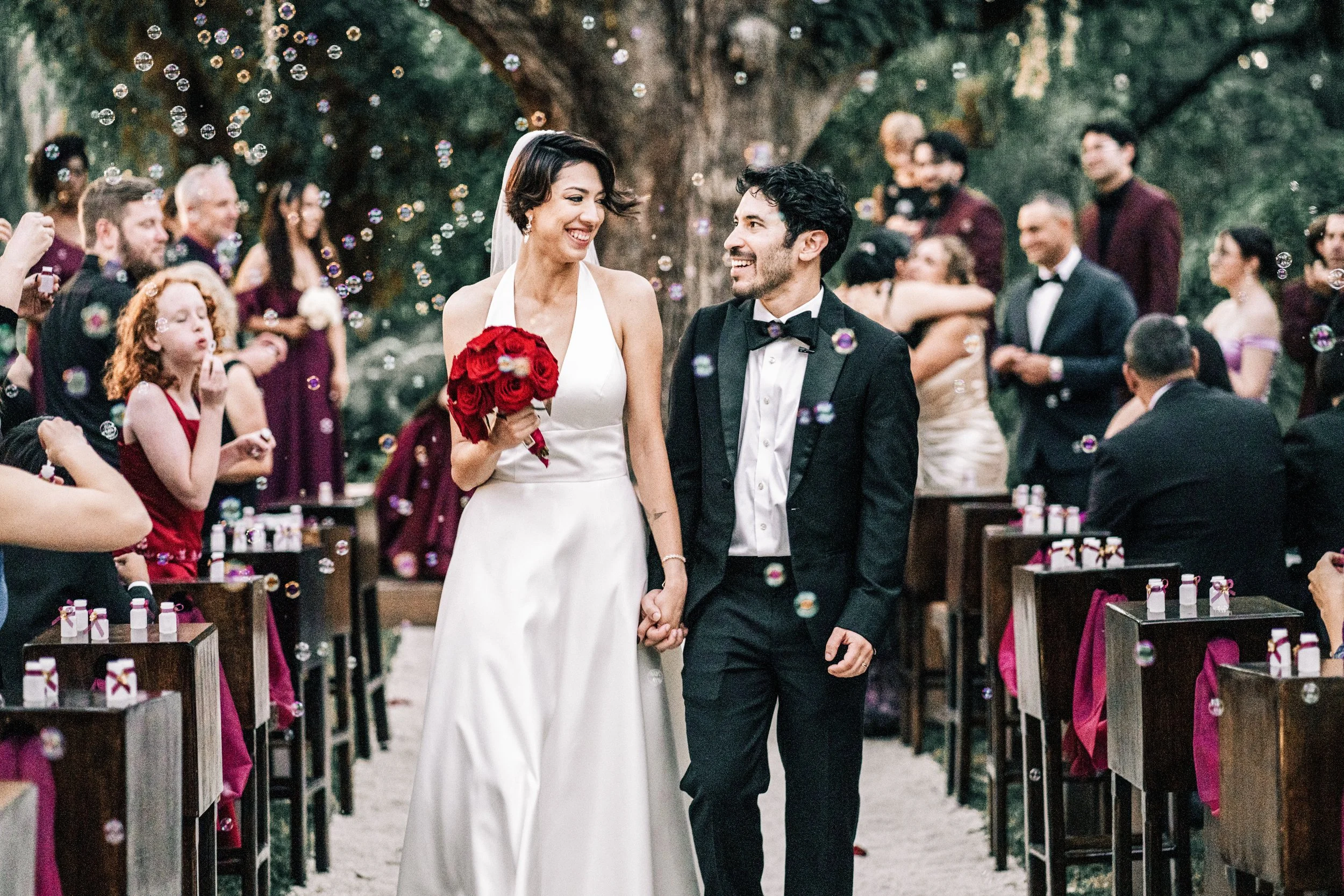 A bride and groom holding hands, smiling, walking down an outdoor wedding aisle surrounded by guests and decorated chairs, with bubbles floating in the air.