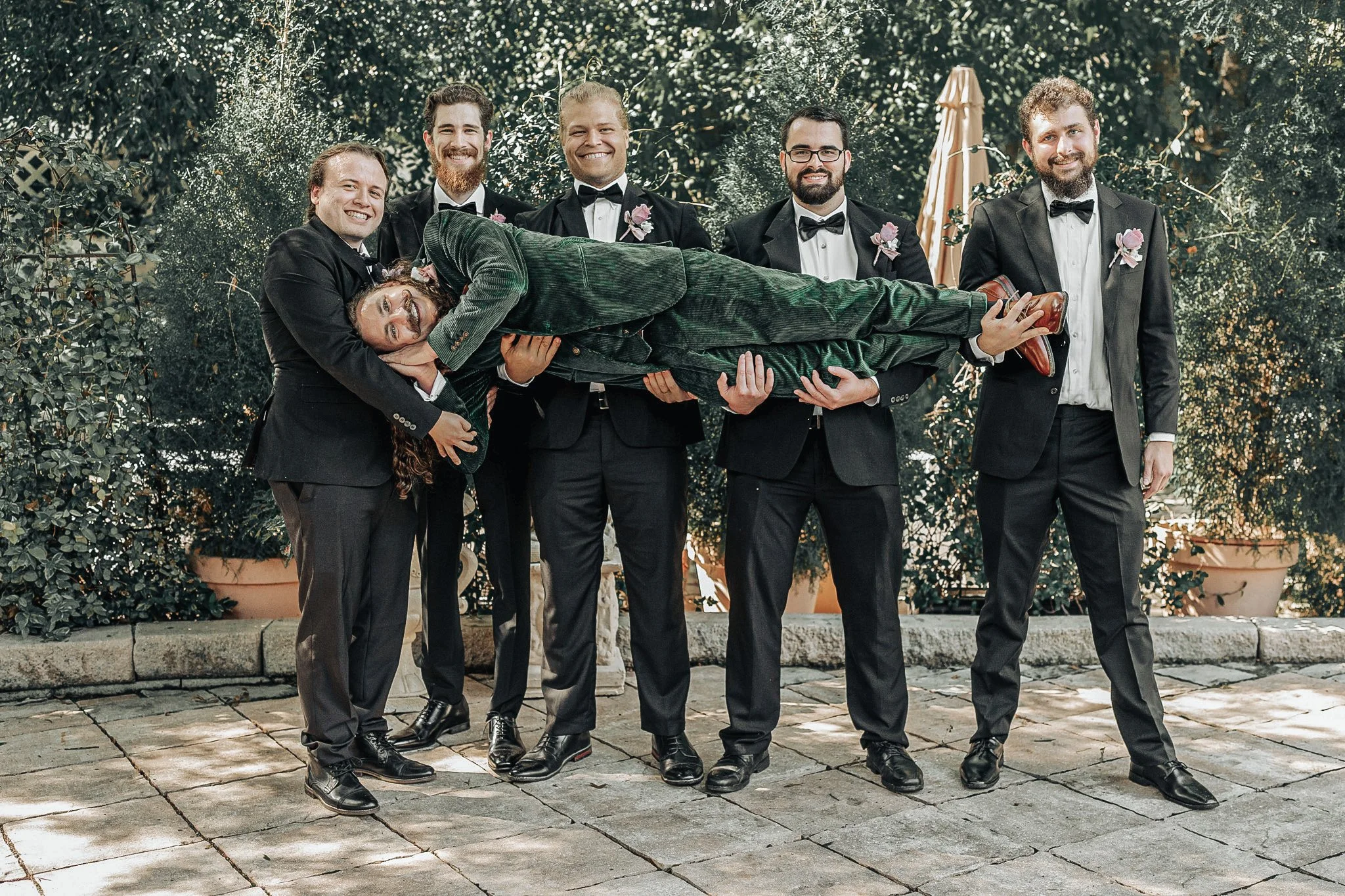 A group of seven men dressed in black tuxedos, with some wearing bow ties, are standing outdoors on a stone-paved area with trees and plants in the background. One man is lying horizontally across the arms of three others, smiling and wearing a green