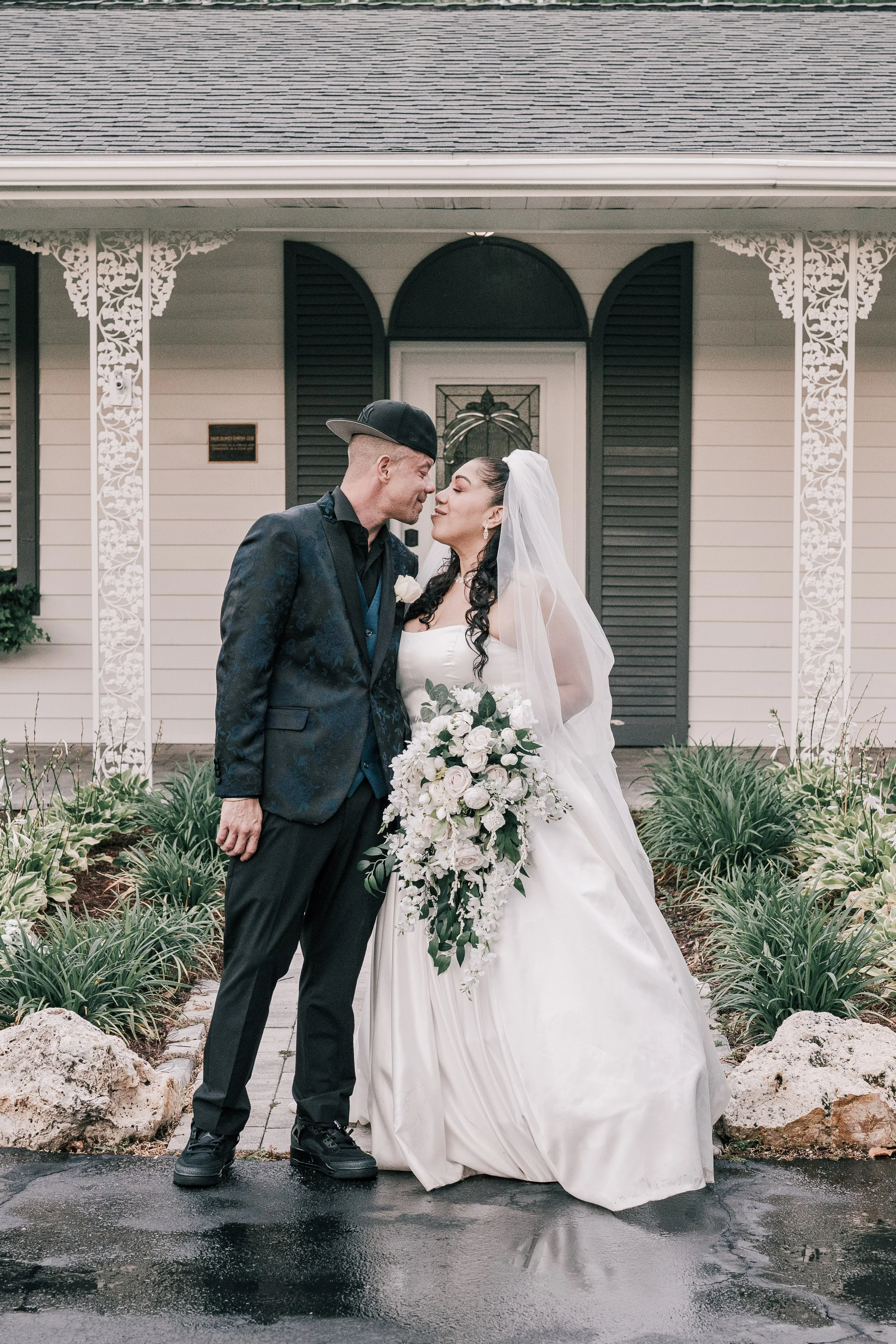 A bride and groom standing close together in front of a house, holding hands and smiling at each other. The bride is wearing a white wedding dress and veil, holding a large bouquet of white flowers. The groom is dressed in a dark suit with a patterne