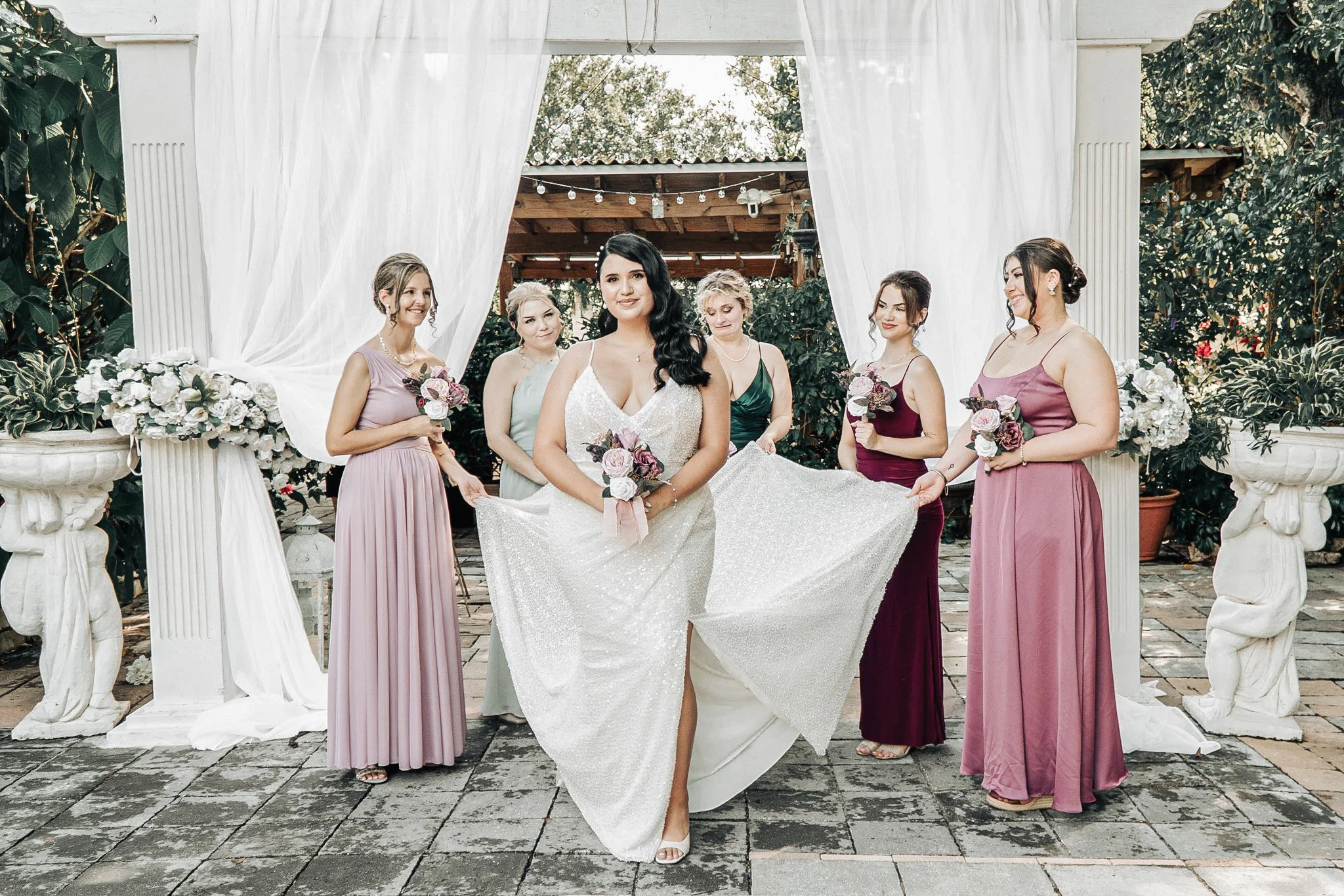 A bride in a white wedding dress holding a bouquet, standing in front of a decorative floral arch with six bridesmaids in pastel and burgundy dresses, outdoors on a stone patio with greenery and string lights in the background.