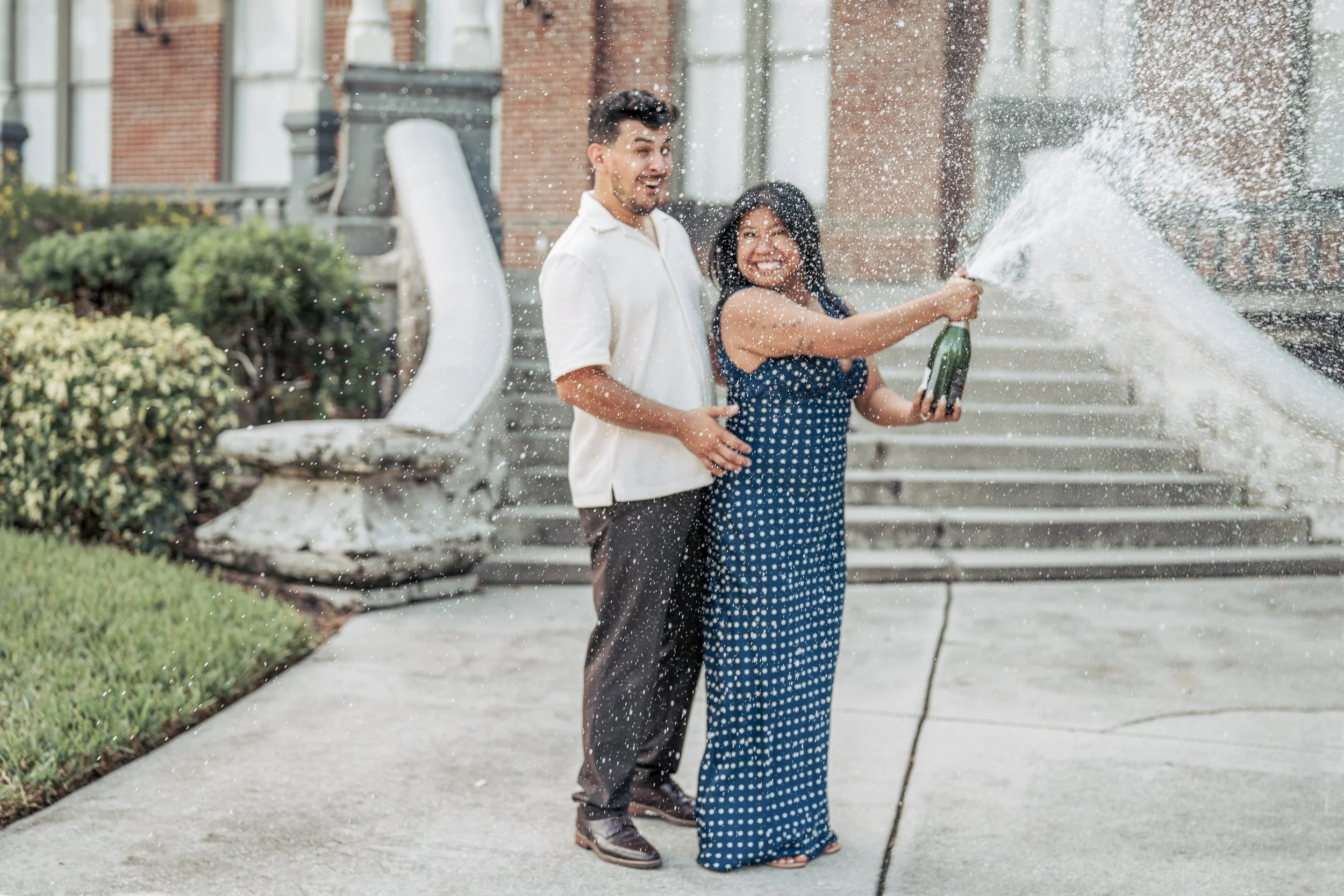 A man and woman are standing on steps outside a building, smiling and holding a champagne bottle as they celebrate, with water spraying from the bottle.