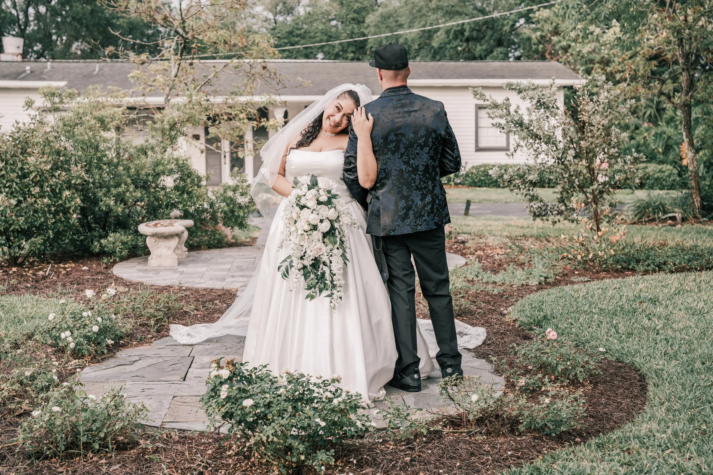A bride and groom stand close together outdoors, smiling, during their wedding. The bride wears a white wedding gown and veil, holding a bouquet of white flowers, while the groom wears a dark suit and hat. There are trees and a house in the backgroun