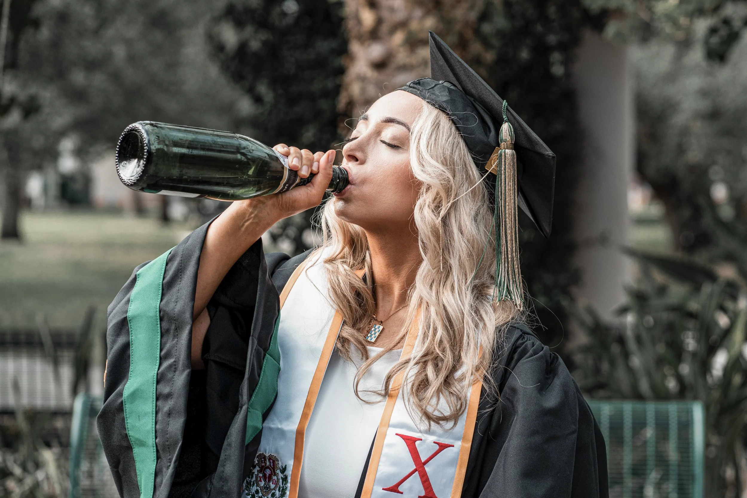 A young woman in graduation attire, including a cap with a tassel, is drinking from a large bottle of a beverage, possibly sparkling water or soda, outdoors in a park setting.