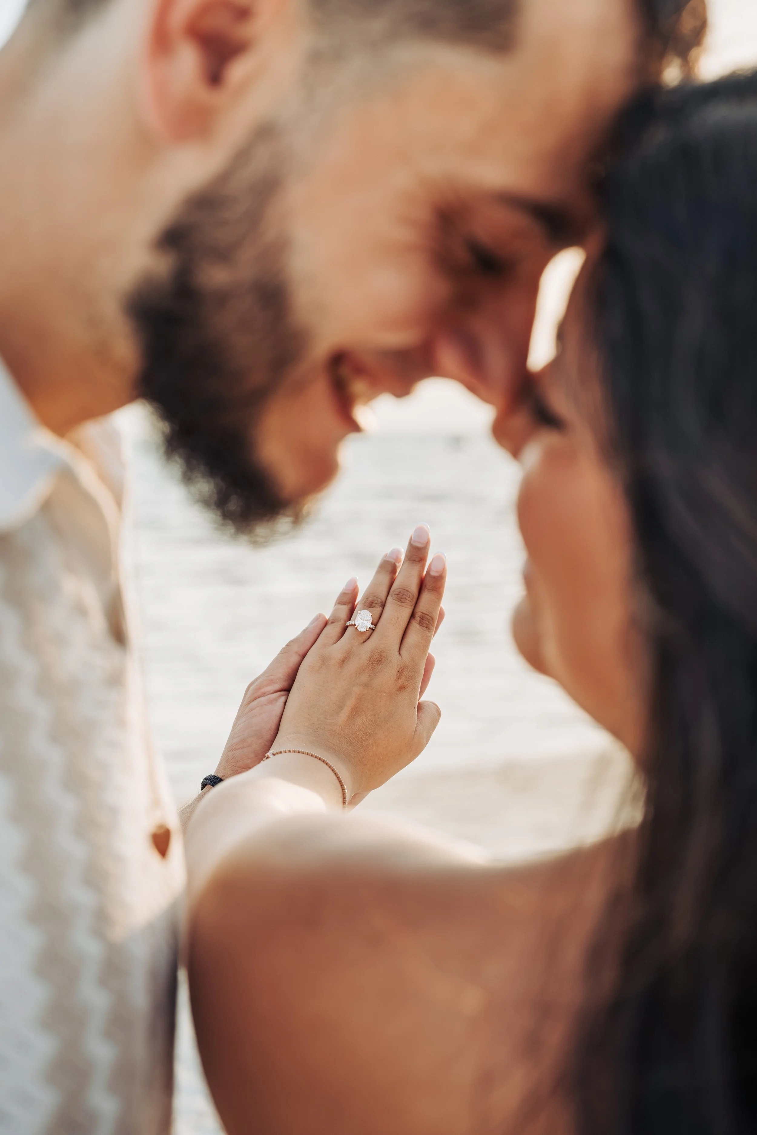 Close-up of a couple touching foreheads at the beach, with a woman's hand showing an engagement ring, symbolizing love and engagement.