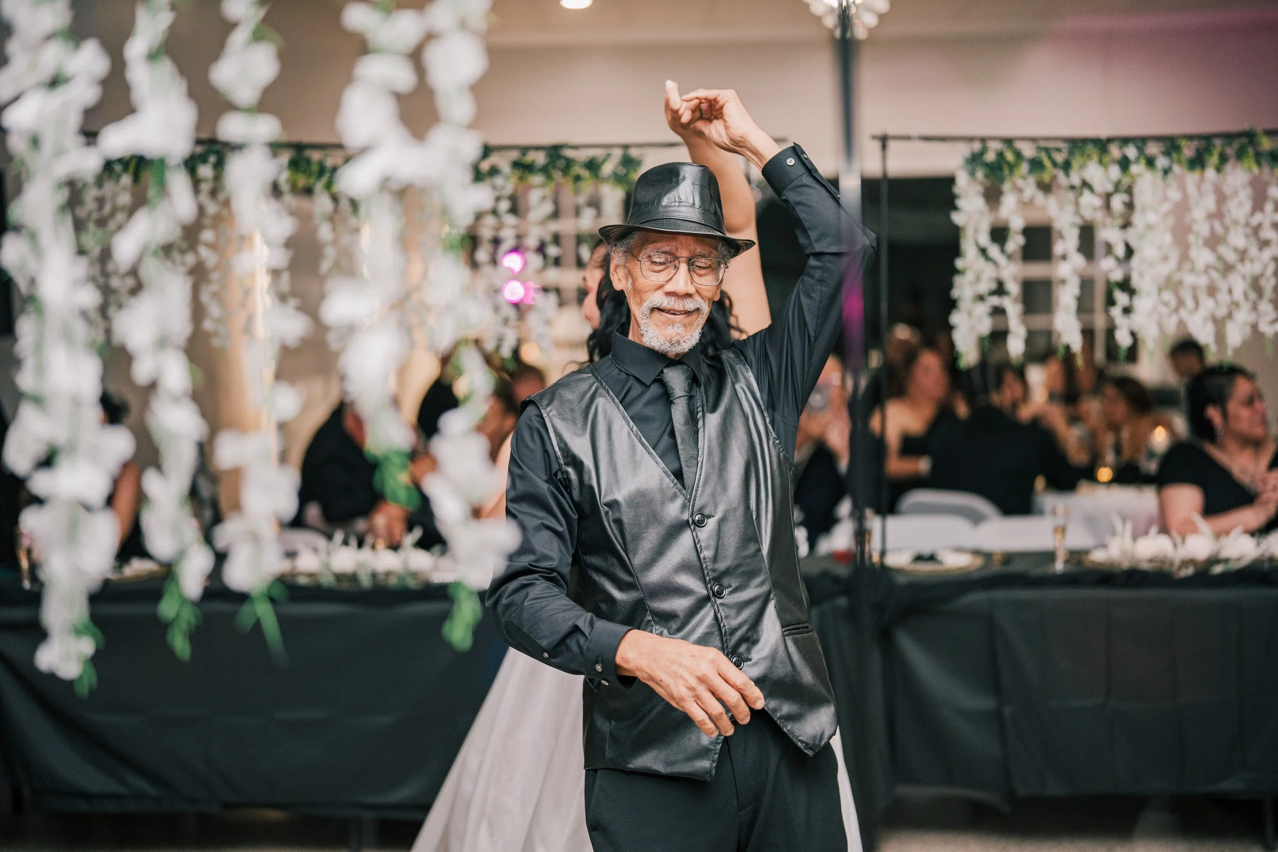 A man with long hair, glasses, and a beard, dressed in a black shiny shirt and a black hat, dancing at a social gathering with hanging white flowers and many seated guests in the background.