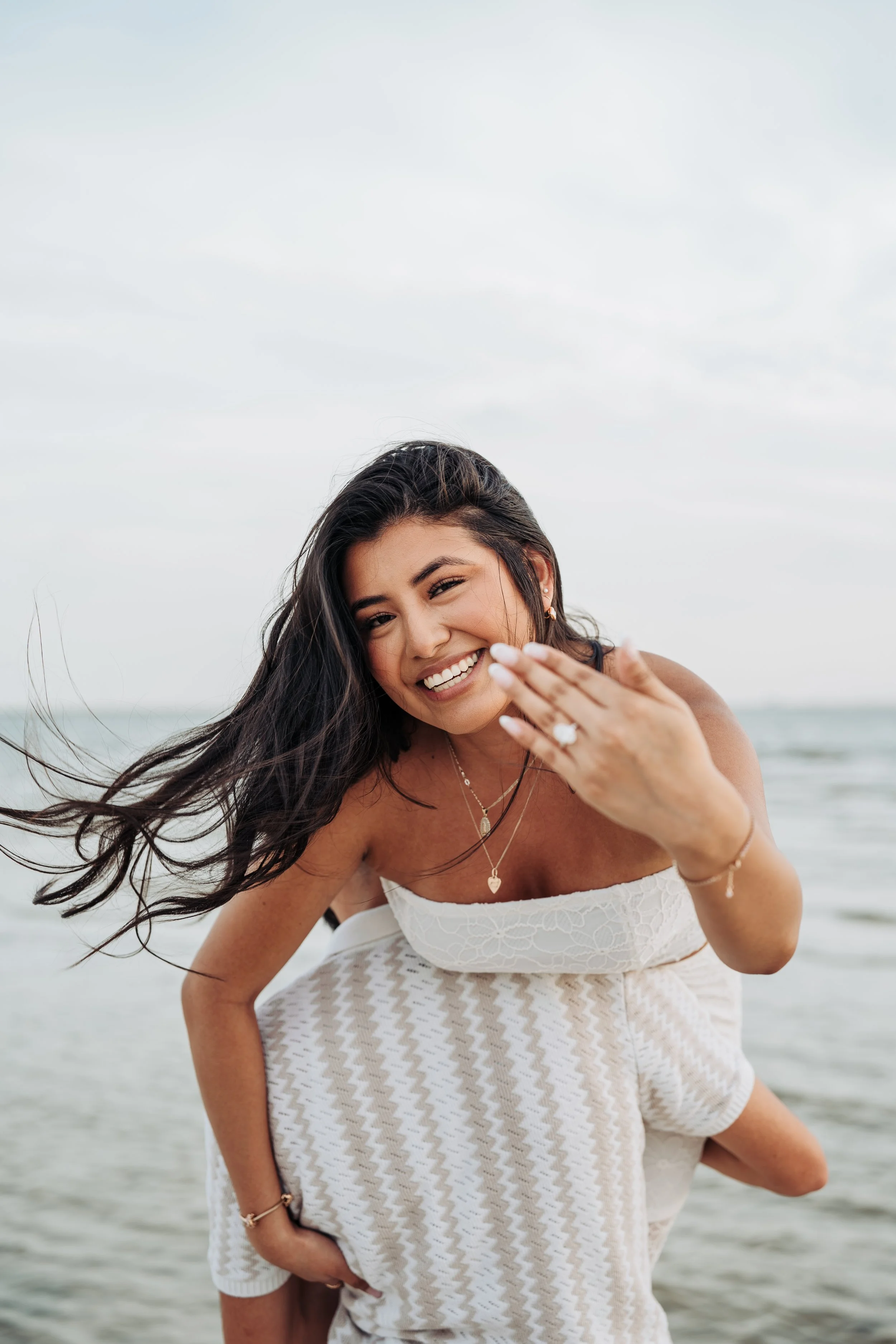 A woman smiling at the camera, standing on a beach with ocean and cloudy sky in the background.
