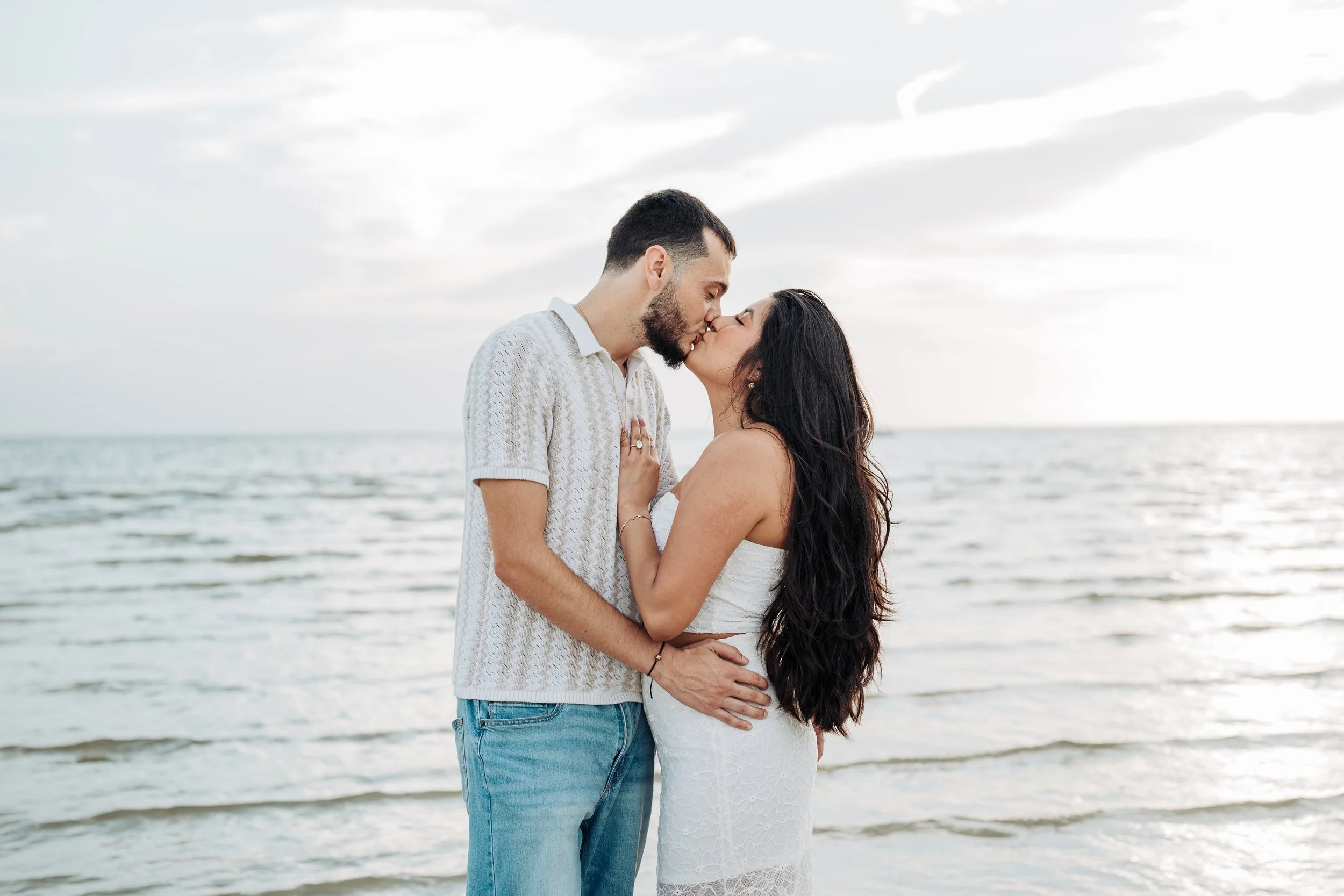 A couple kissing on a beach with the ocean in the background, the sky partly cloudy.