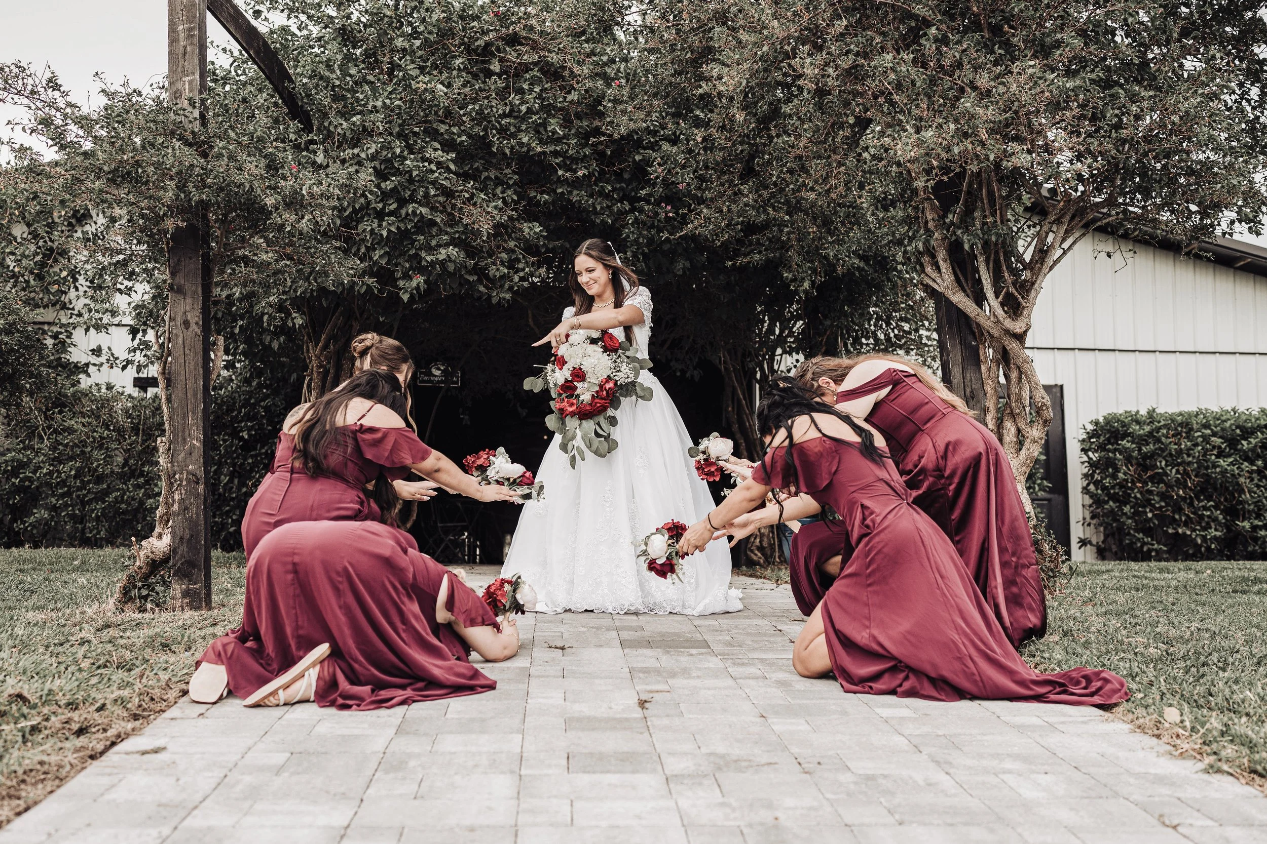A bride in a white wedding dress holding a bouquet of flowers standing on a pathway under a large tree during her wedding ceremony. Bridesmaids in maroon dresses are kneeling on the ground, holding flowers and bowing.