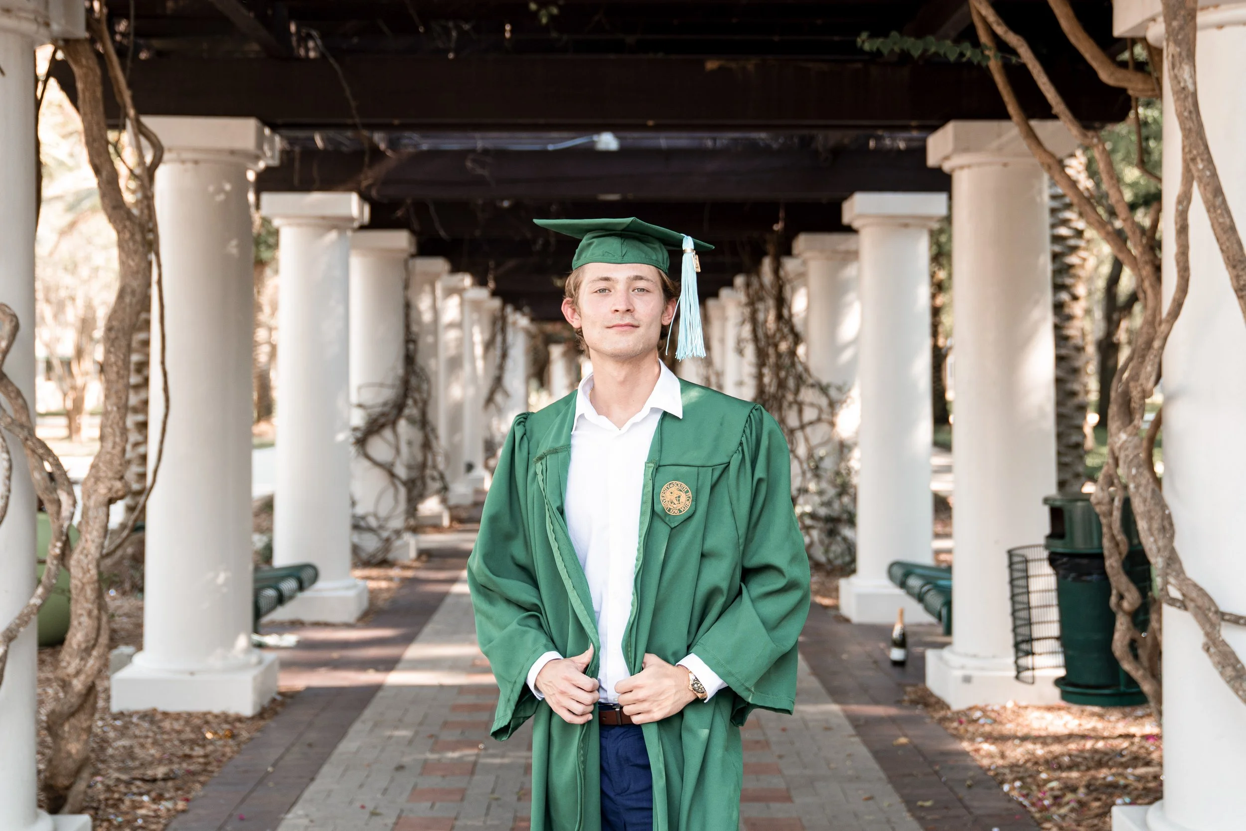 A young man in a green graduation gown and cap standing outdoors under a pergola with white columns and twisted tree branches.