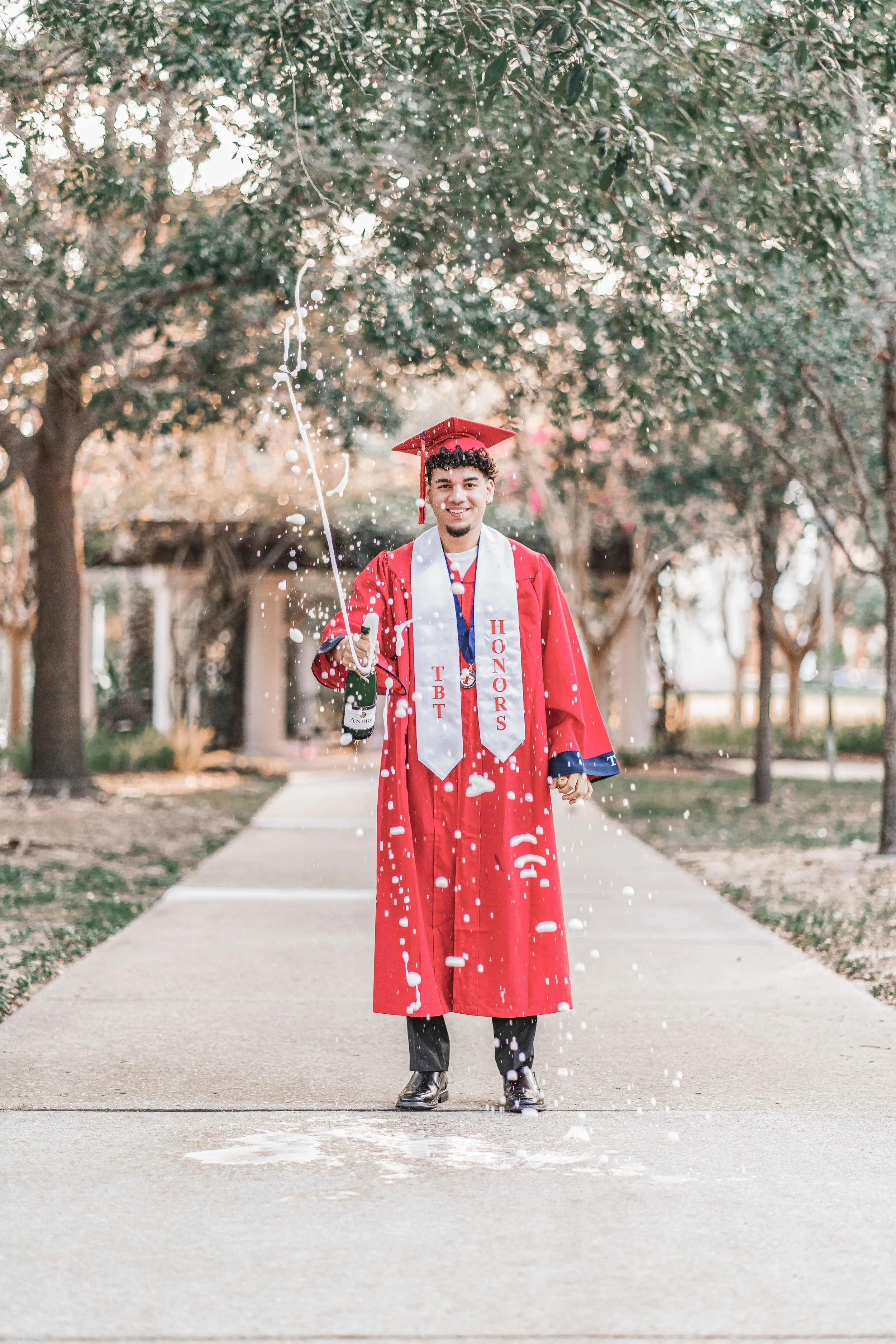 A young man in a red graduation gown and mortarboard holding a bottle of champagne and celebrating on a sidewalk with trees in the background. He is smiling and spraying champagne, with some foam on his shoes, during daytime.