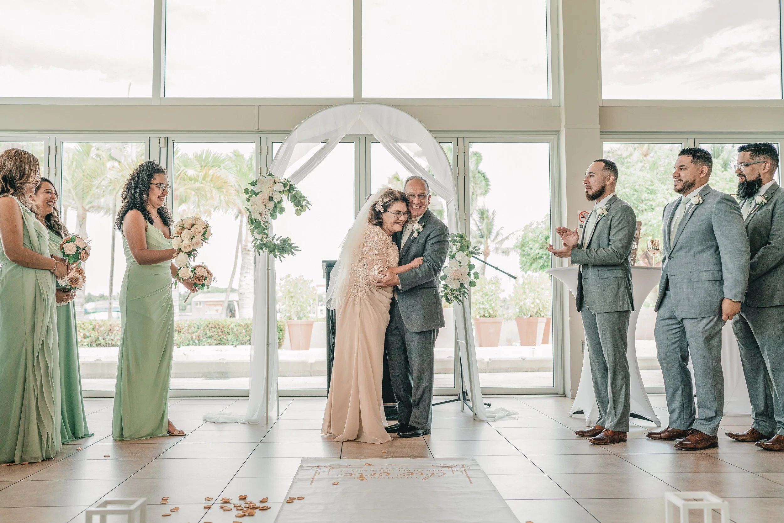 A wedding ceremony in a bright indoor space with large windows and palm trees outside. An older woman in an ivory dress and an older man in a gray suit are hugging and smiling at the center. Four women in matching light green dresses holding bouquets