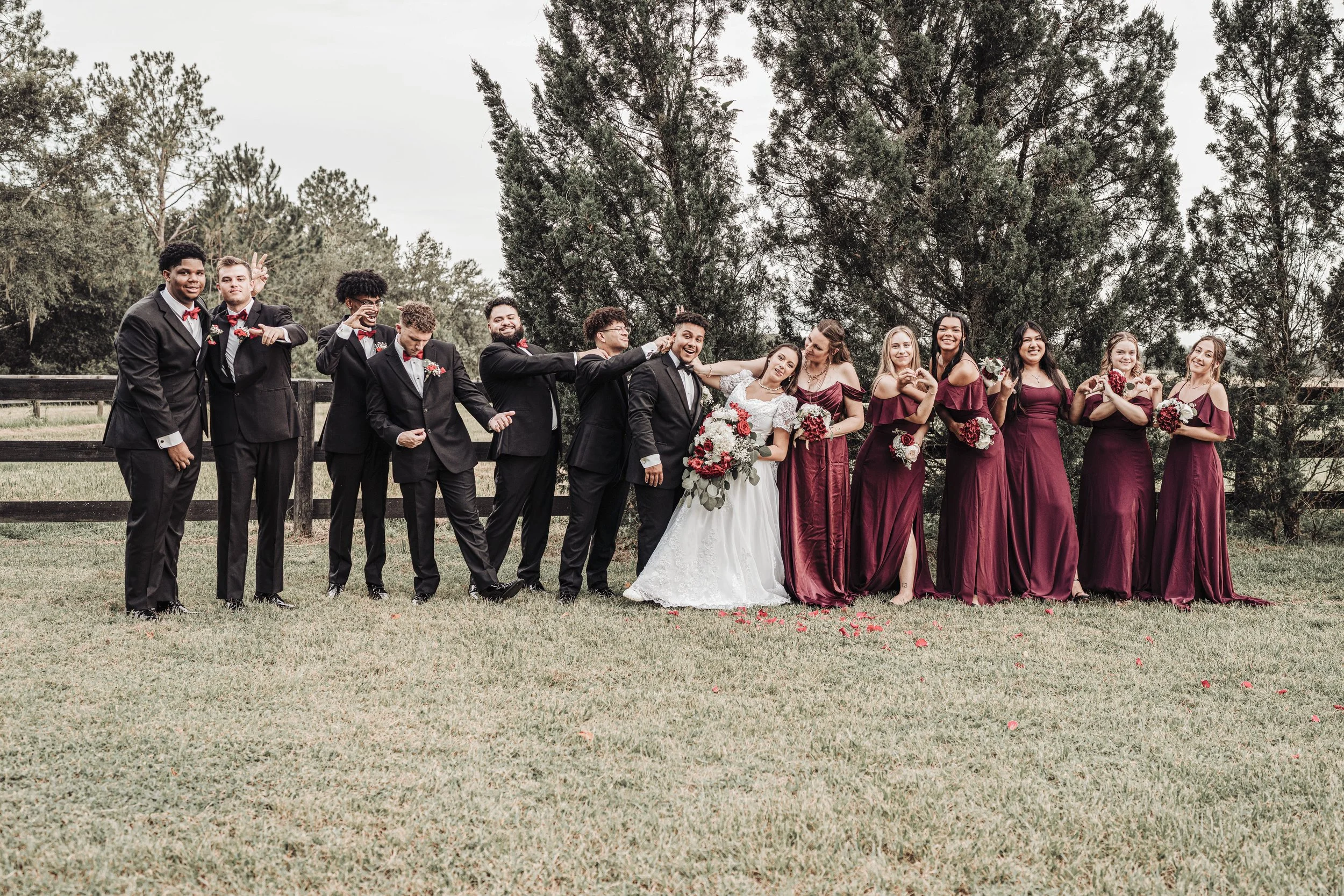 A wedding party of eleven people, six men and five women, standing outdoors on grass with trees in the background. The bride and groom are in the center, with the bride holding a bouquet and the groom in a tuxedo. The women are dressed in burgundy dr