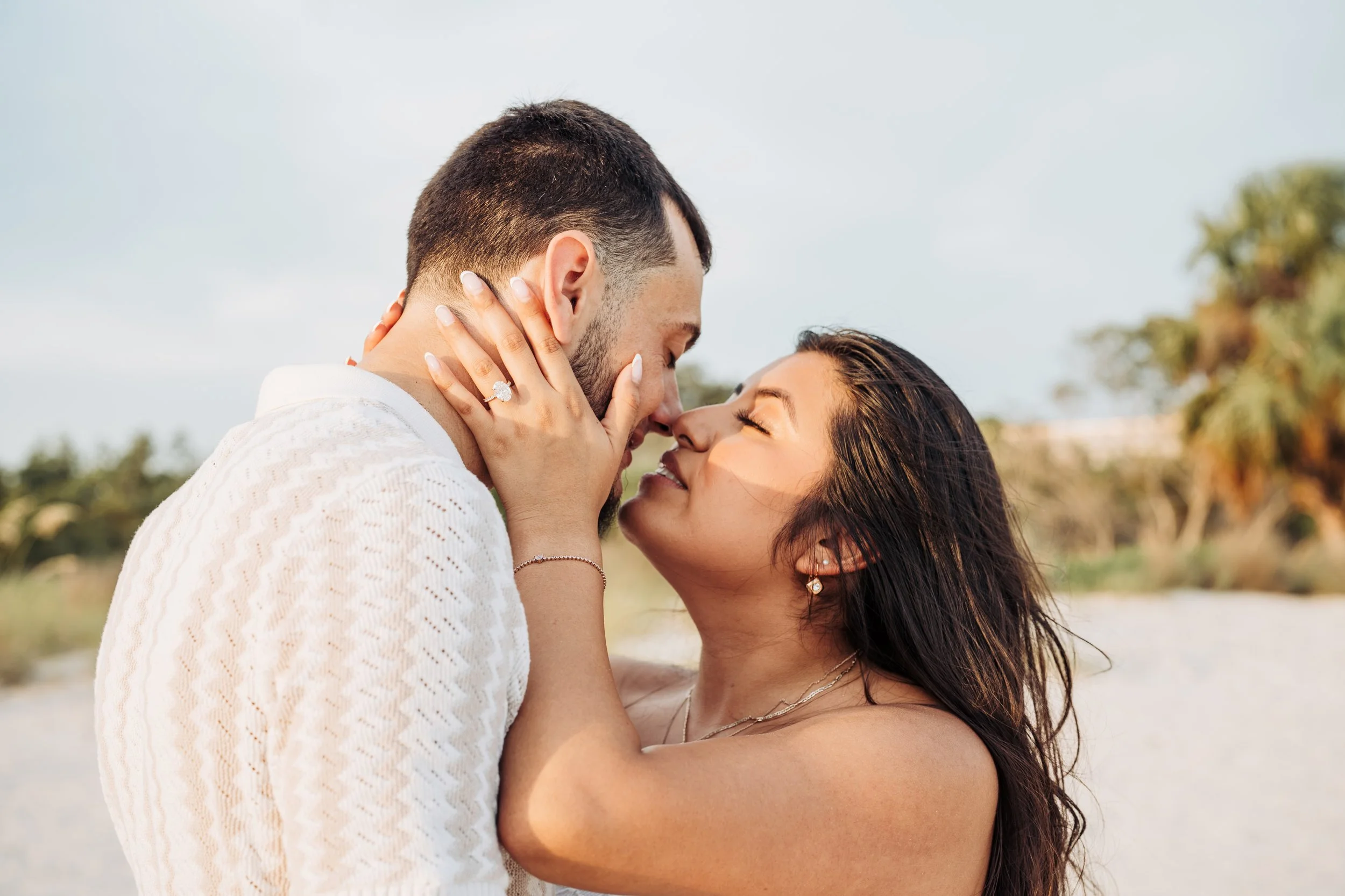 A couple sharing a romantic moment outdoors, nose to nose, with the woman holding the man's face, both smiling with eyes closed, in a natural setting with trees and sky in the background.