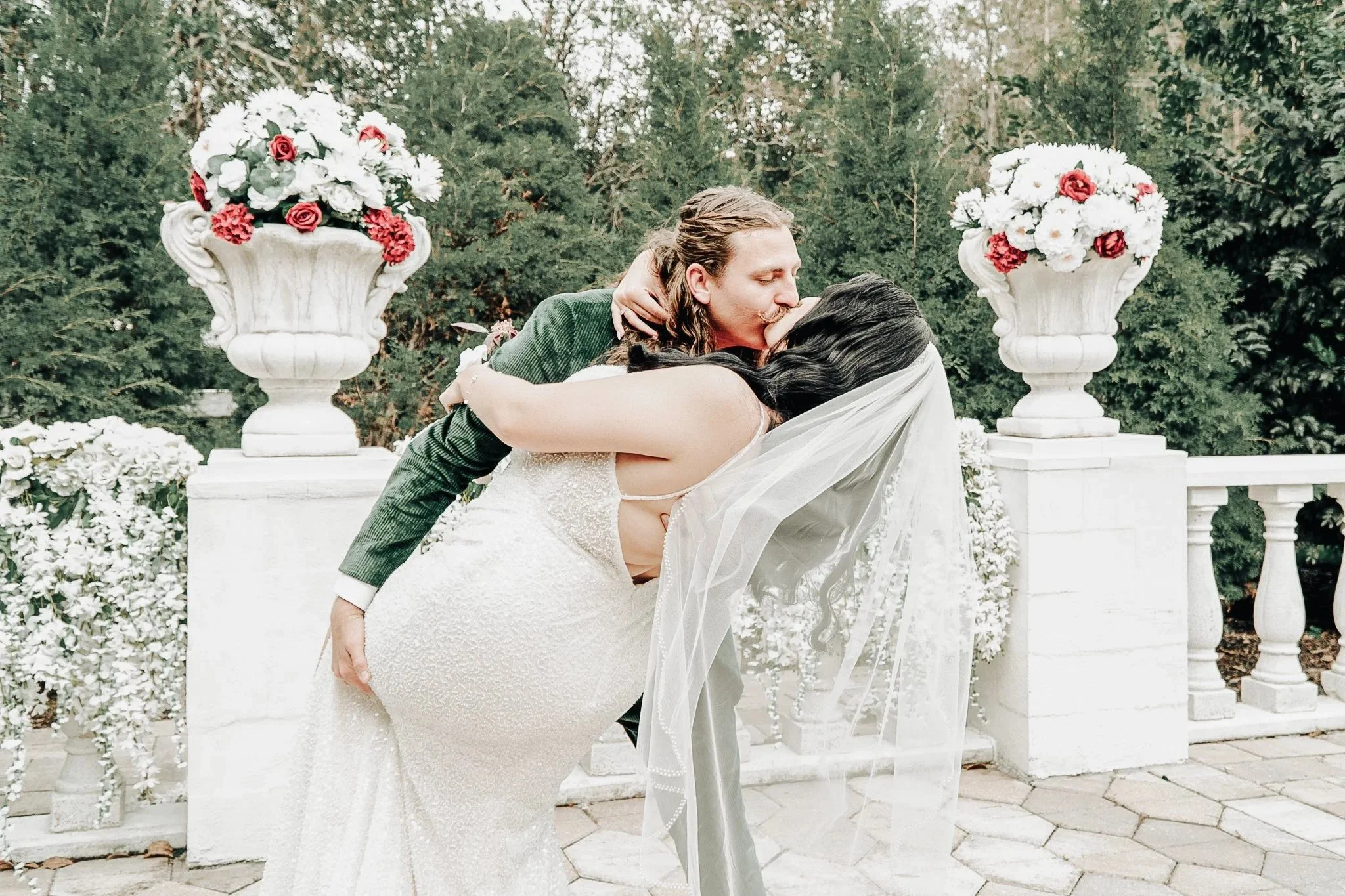 A man and woman are kissing outdoors during a wedding ceremony, with the man wearing a green jacket and the woman in a white wedding dress. They are surrounded by white flowers and greenery.