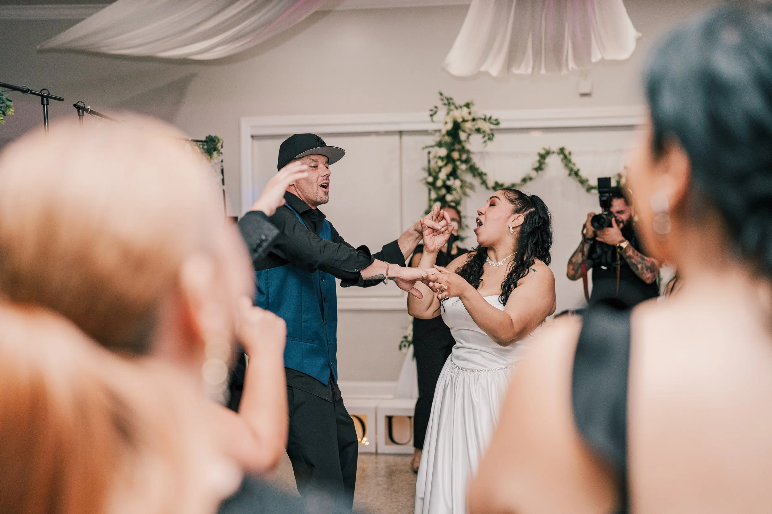 A bride and groom dancing at their wedding with guests in the background, one guest holding a camera and a decorative floral arrangement on the wall.