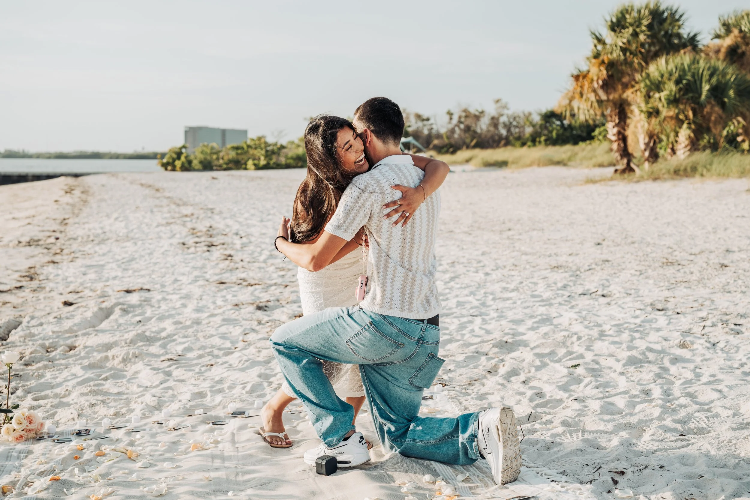 A woman and man embrace on a sandy beach during a wedding ceremony, with candles and roses on a cloth-covered table nearby, palm trees in the background, sunlight, and a calm body of water.