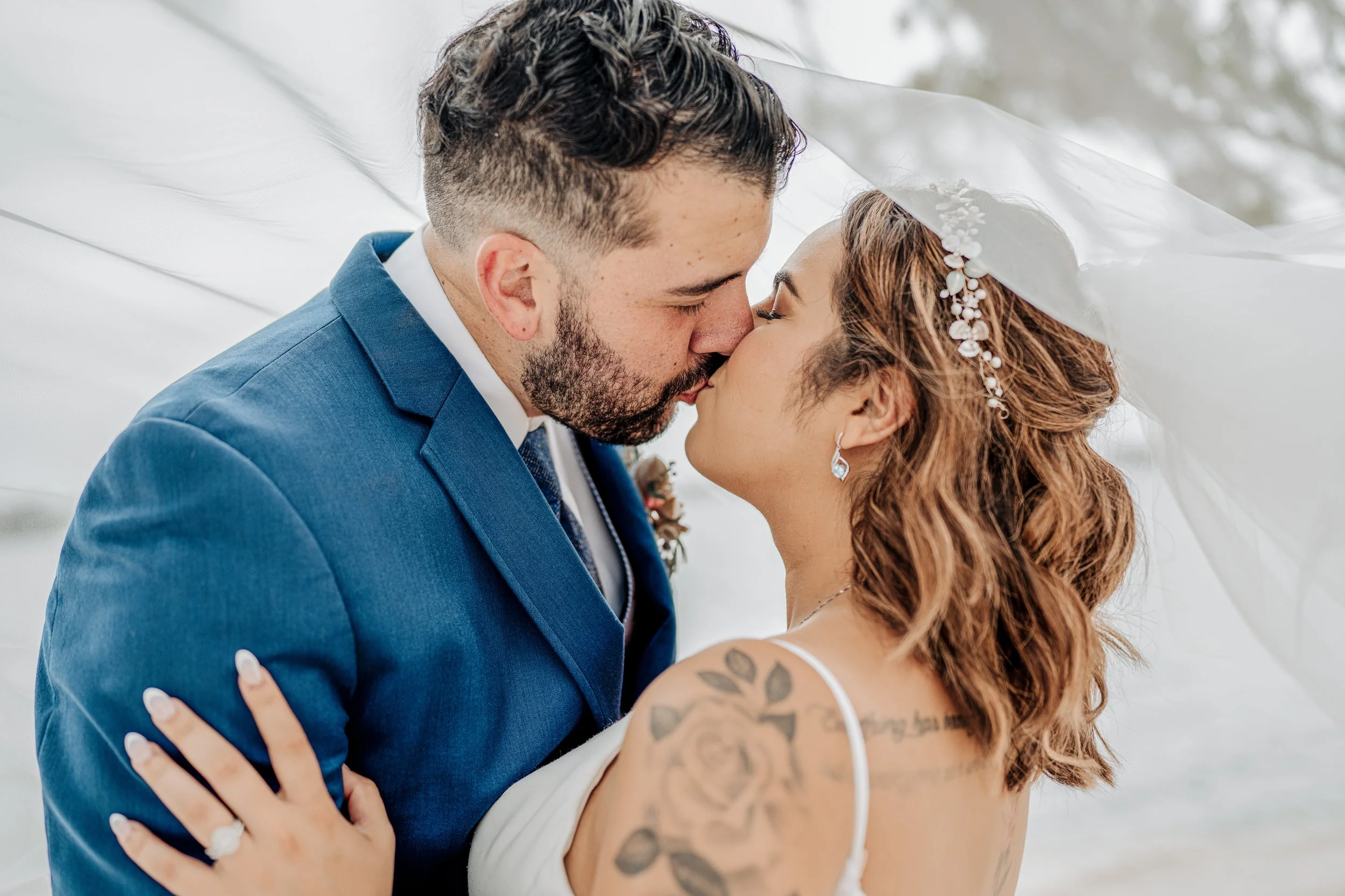 A bride and groom share a kiss under a white veil at their wedding ceremony, with the groom wearing a blue suit and the bride with tattoos on her arms and wearing a floral hairpiece.