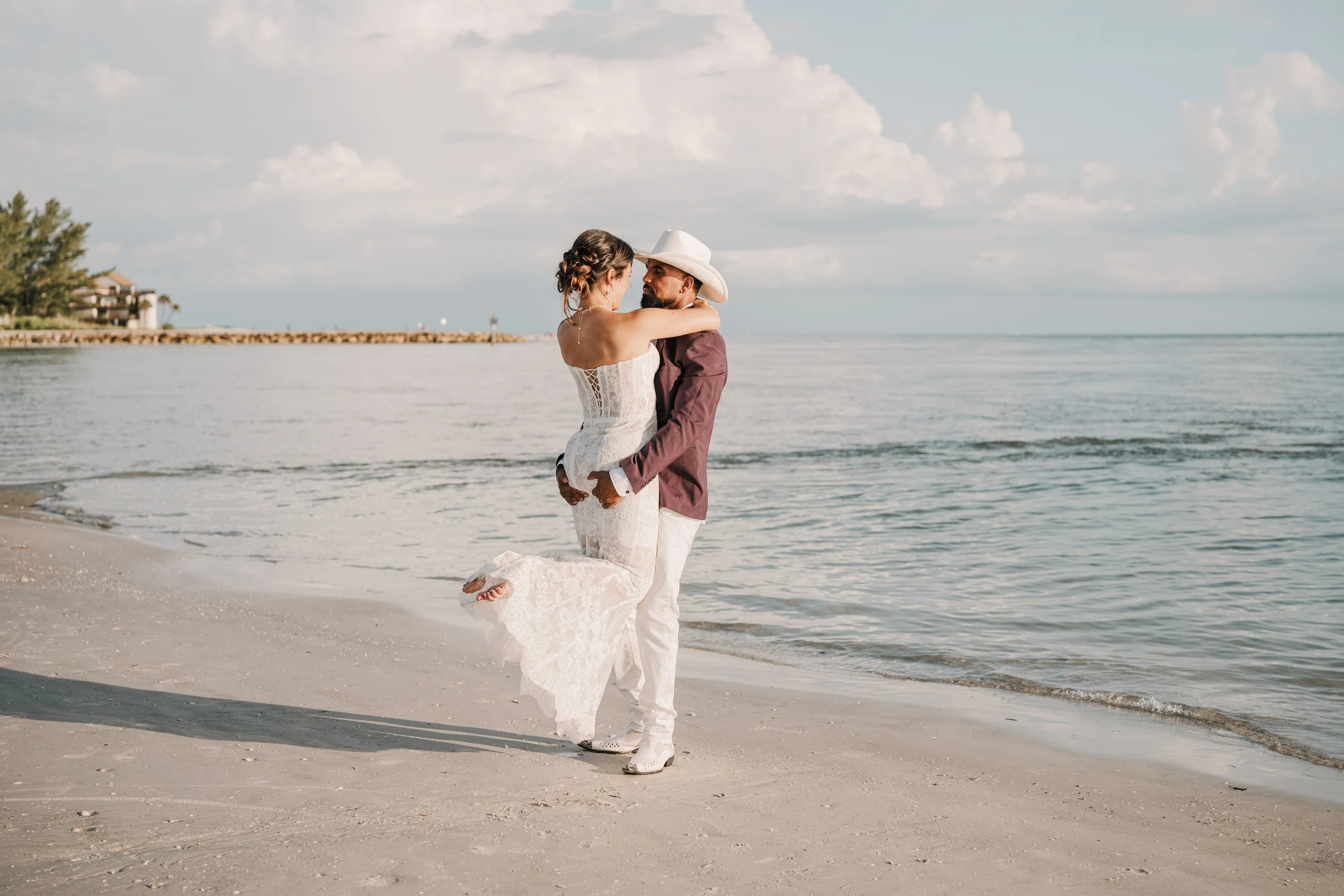 A couple dressed in wedding attire on the beach, with the man lifting the woman in his arms. The woman is wearing a white lace wedding dress, and the man is wearing a maroon suit, white pants, and a cowboy hat. The ocean and a partly cloudy sky are i