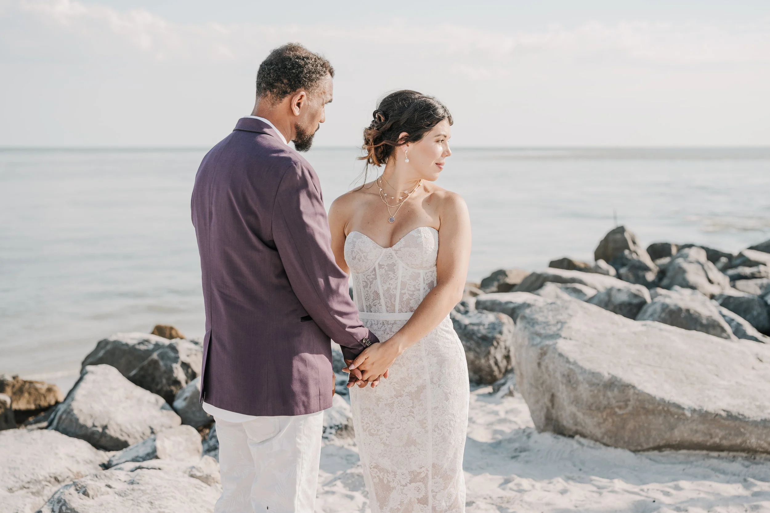 A couple dressed in wedding attire holding hands on a rocky beach with ocean waves in the background.