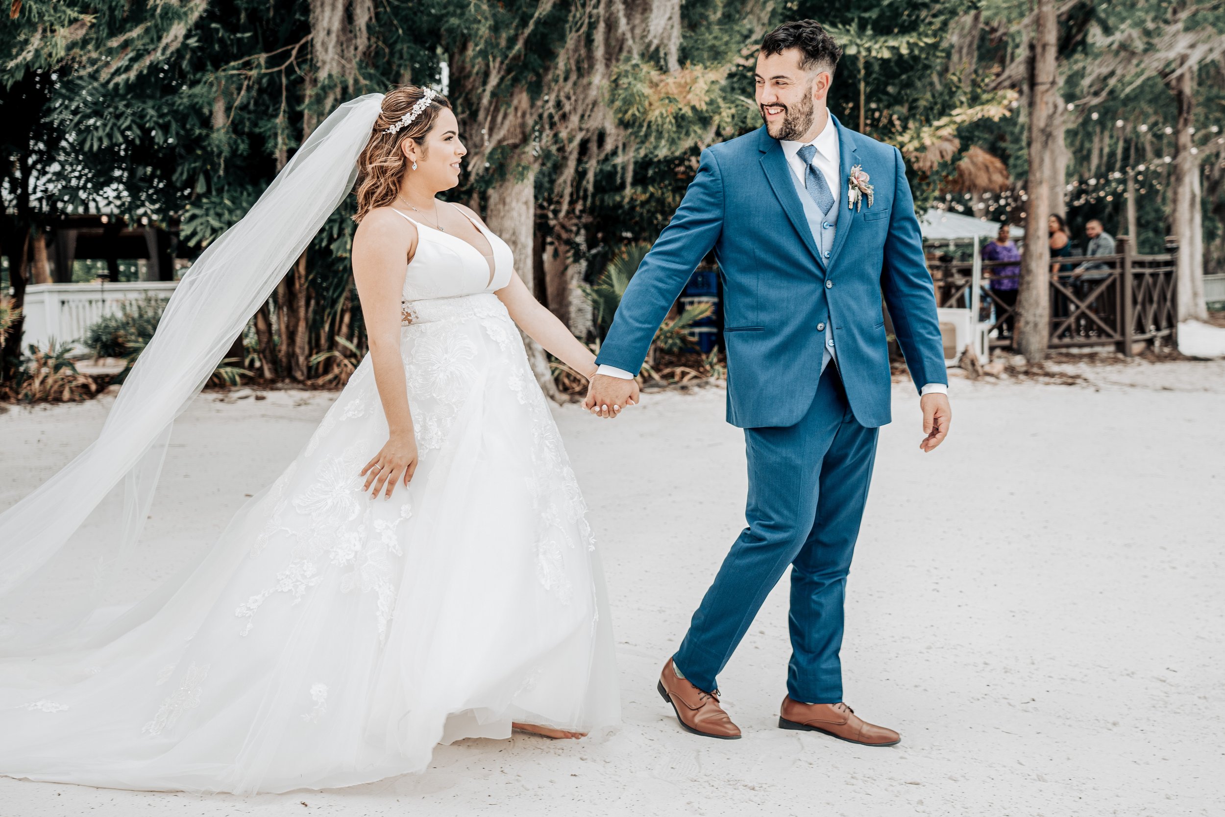 A bride and groom holding hands and smiling during their outdoor wedding ceremony on a sandy area surrounded by trees. The bride is wearing a white wedding gown with lace details and a veil, while the groom is dressed in a blue suit with a patterned 