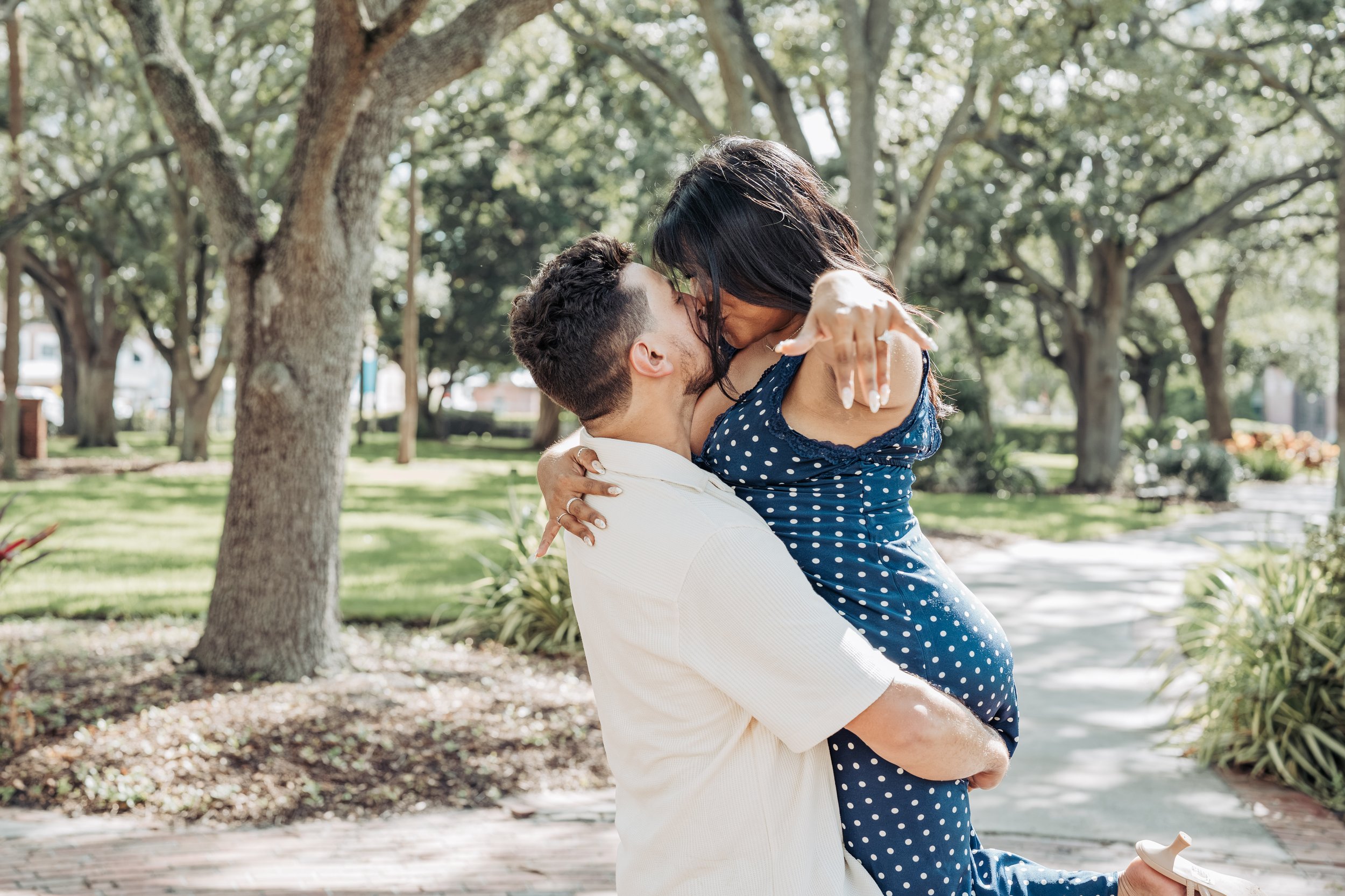 A couple sharing a kiss outdoors in a park, with the woman being lifted off the ground by the man.