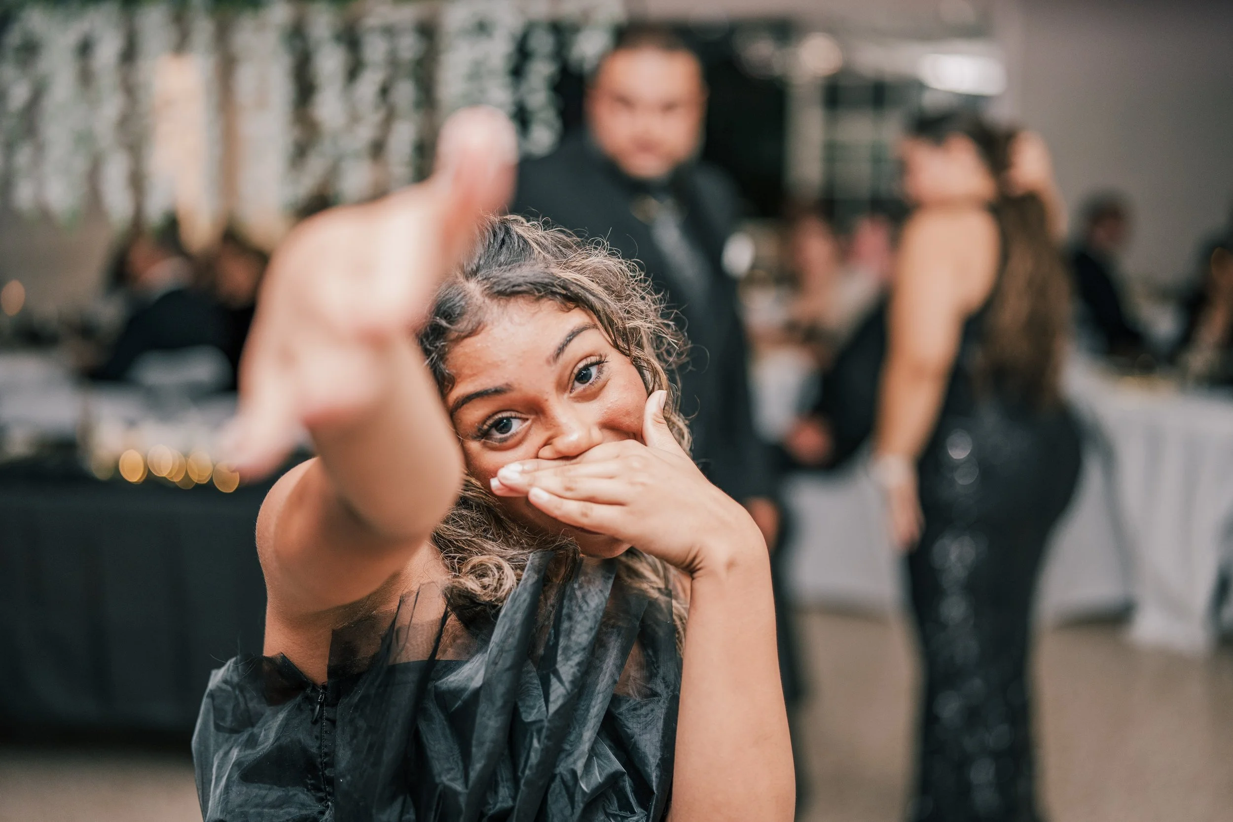 A woman with curly hair covering her mouth with her hand, giving a thumbs-up, at a social event with people in the background.