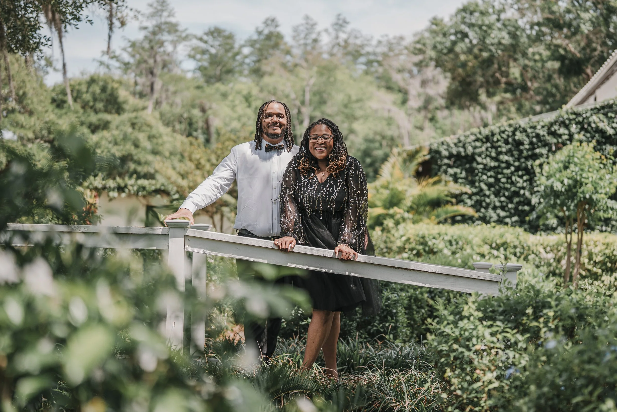 A couple standing on a white outdoor bridge in a lush garden, smiling at the camera, dressed in formal attire with the man in a white shirt and bow tie and the woman in a black dress with sparkle details.