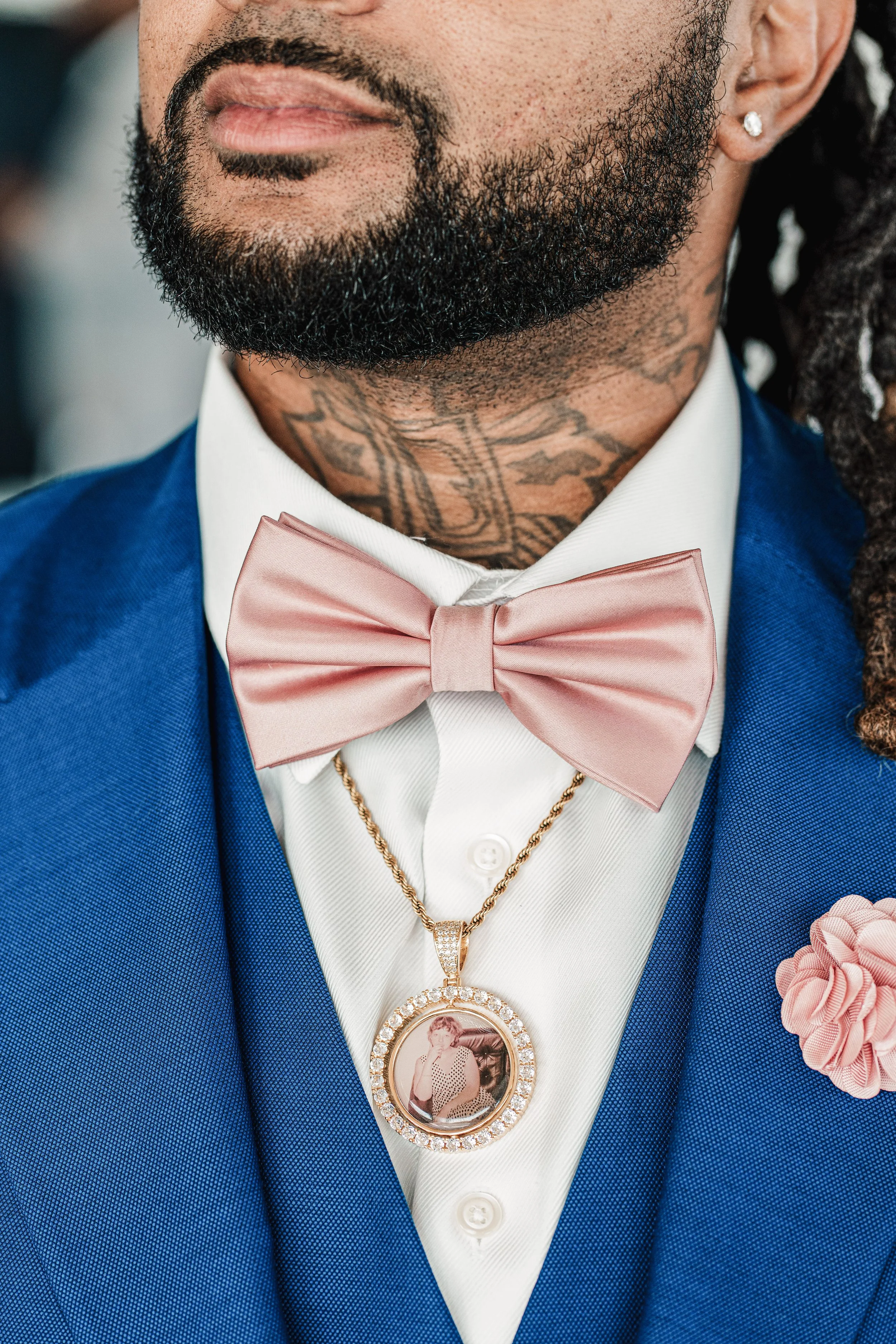 Close-up of a man dressed formally in a blue suit, white shirt, and pink bowtie, wearing jewelry including a gold chain with a pendant, an earring, and a tattoo on the neck. The pendant features a photo of a woman.