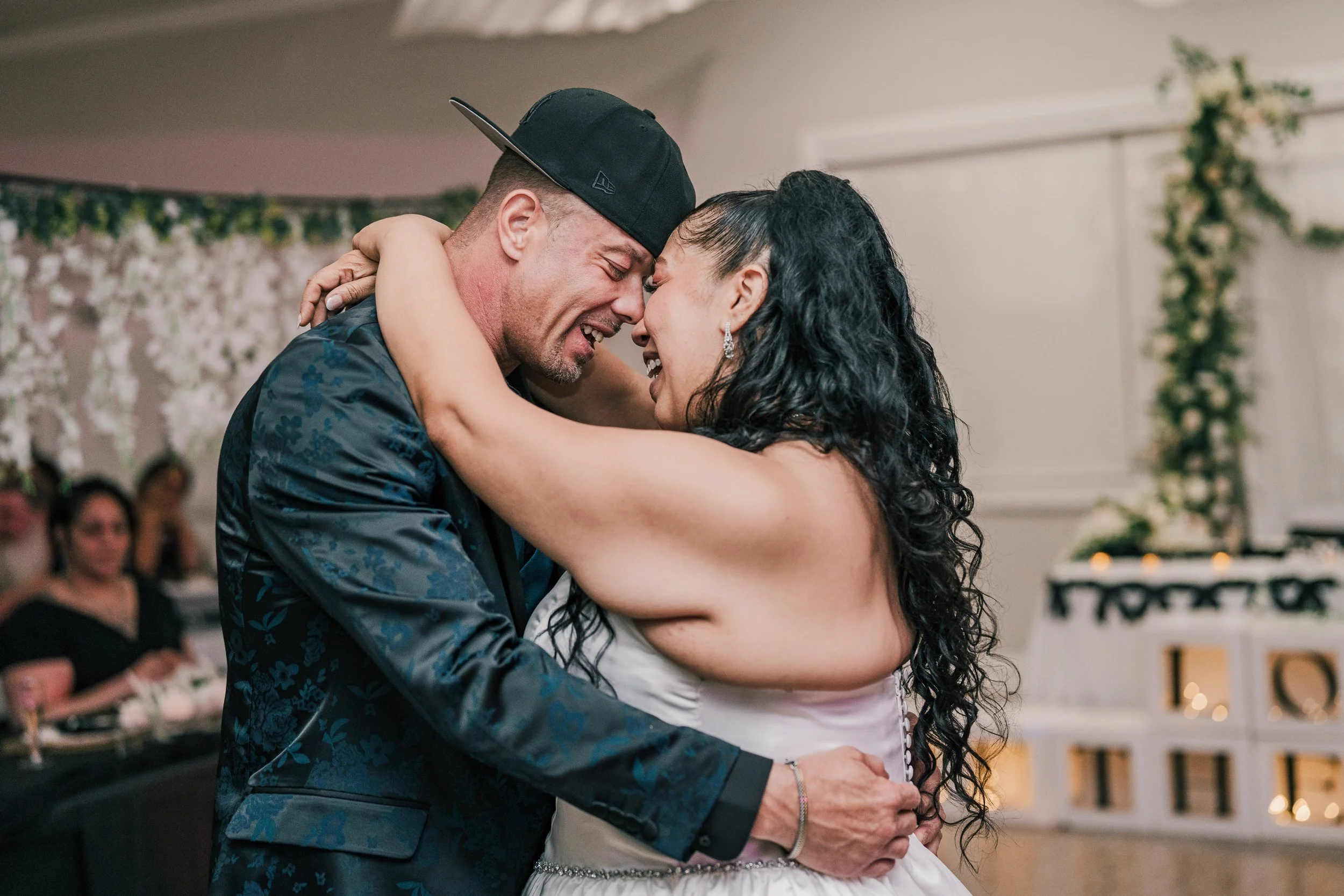 A couple, a man and a woman, share an intimate dance at a wedding reception, smiling with their foreheads touching. The man is wearing a black cap backward and a patterned shirt, and the woman has long dark curly hair and jewelry. In the background, 
