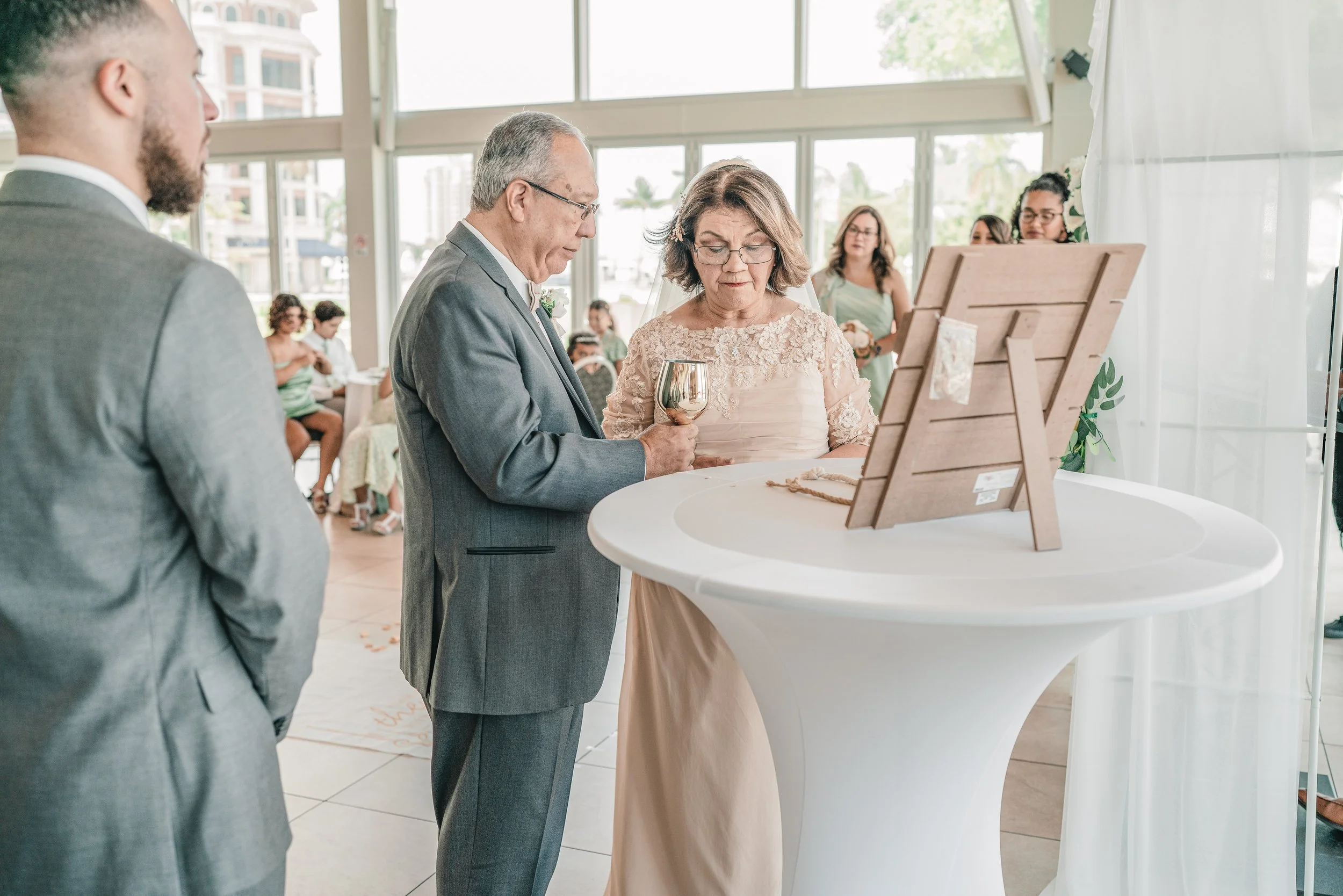 A wedding ceremony inside a bright, modern venue with large windows. An older woman in a lace dress holding a wine glass stands next to a man in a gray suit, both looking at a display. A man in a gray tuxedo is visible on the left, and several guests
