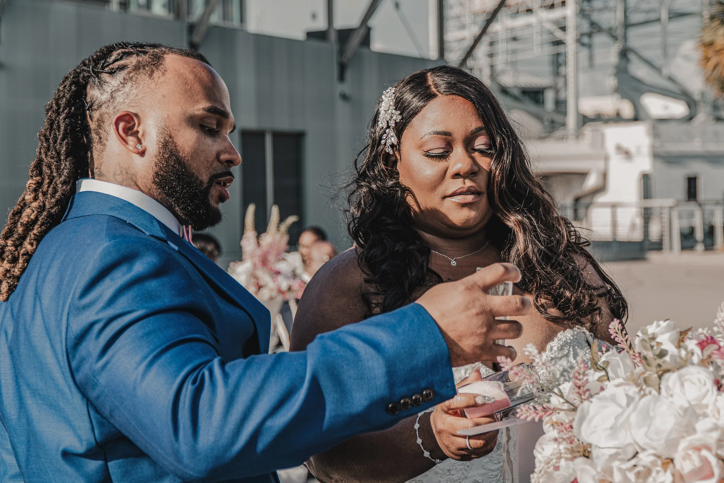 A bride and groom at a wedding look at their phones while standing near a large floral arrangement outdoors, with industrial buildings in the background.