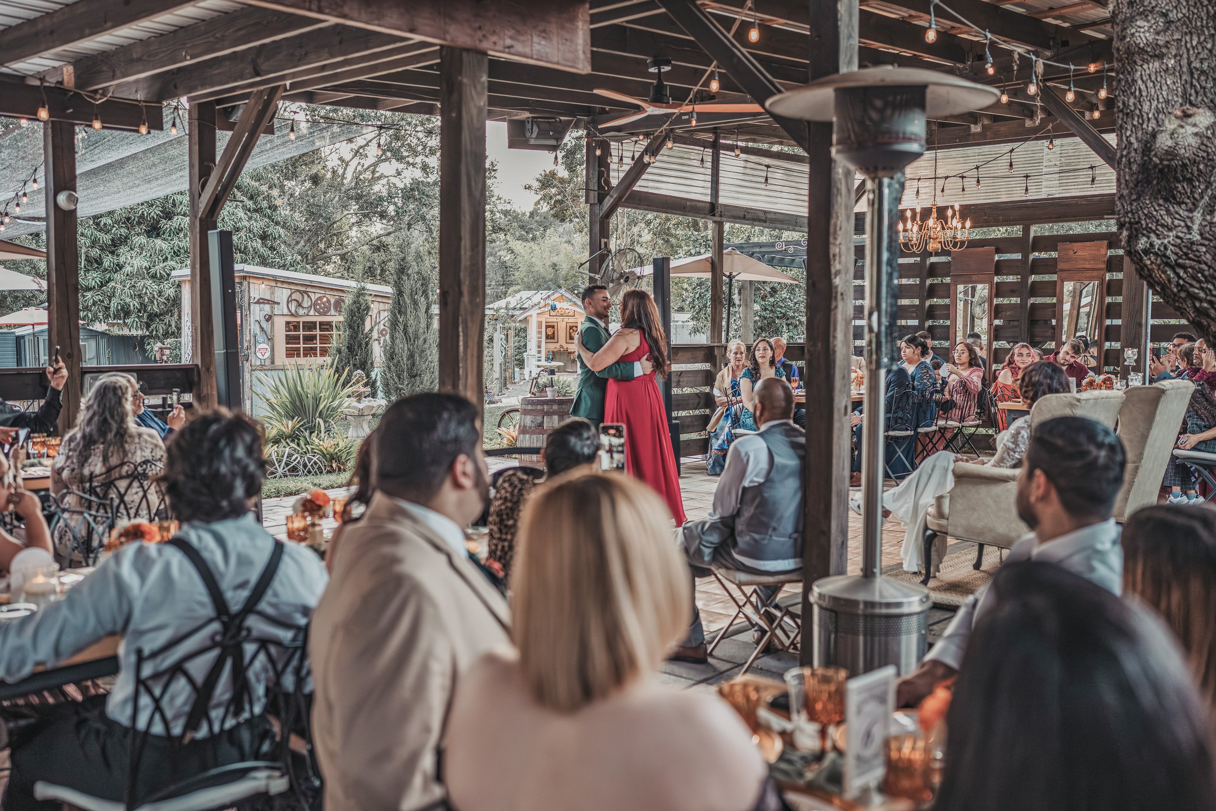 Wedding reception with guests seated at tables and a couple dancing in the center under a rustic outdoor pavilion decorated with string lights, chandeliers, and surrounded by trees.