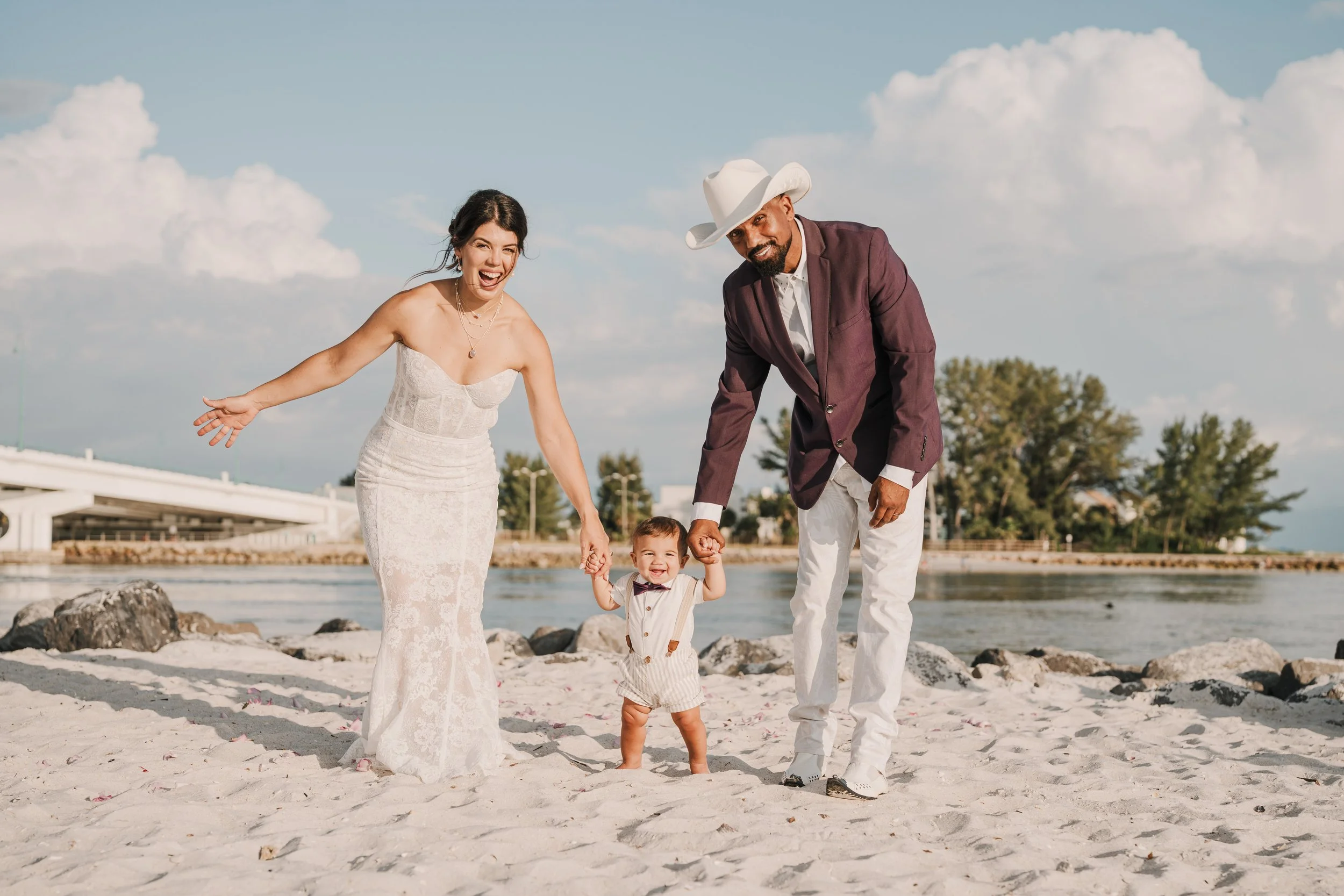 A family of four with light skin enjoying a day at the beach, holding hands and smiling, with a bridge, trees, and clouds in the background.