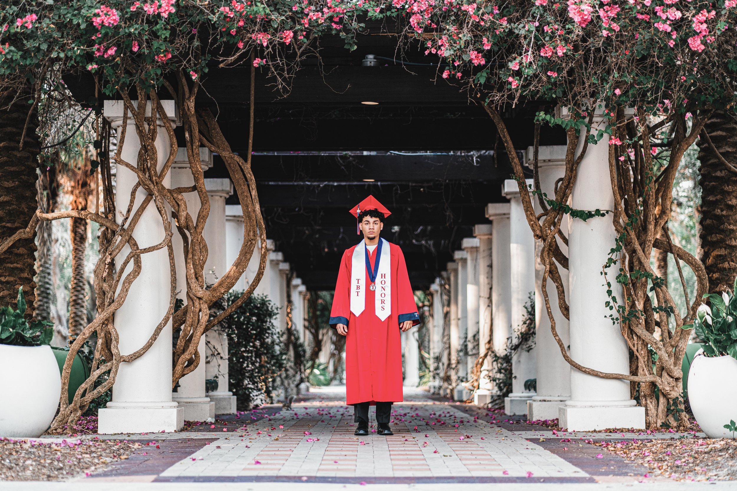 A young man in a red graduation gown and cap standing under a pergola covered with climbing plants and pink flowers, with white columns and potted plants on either side.