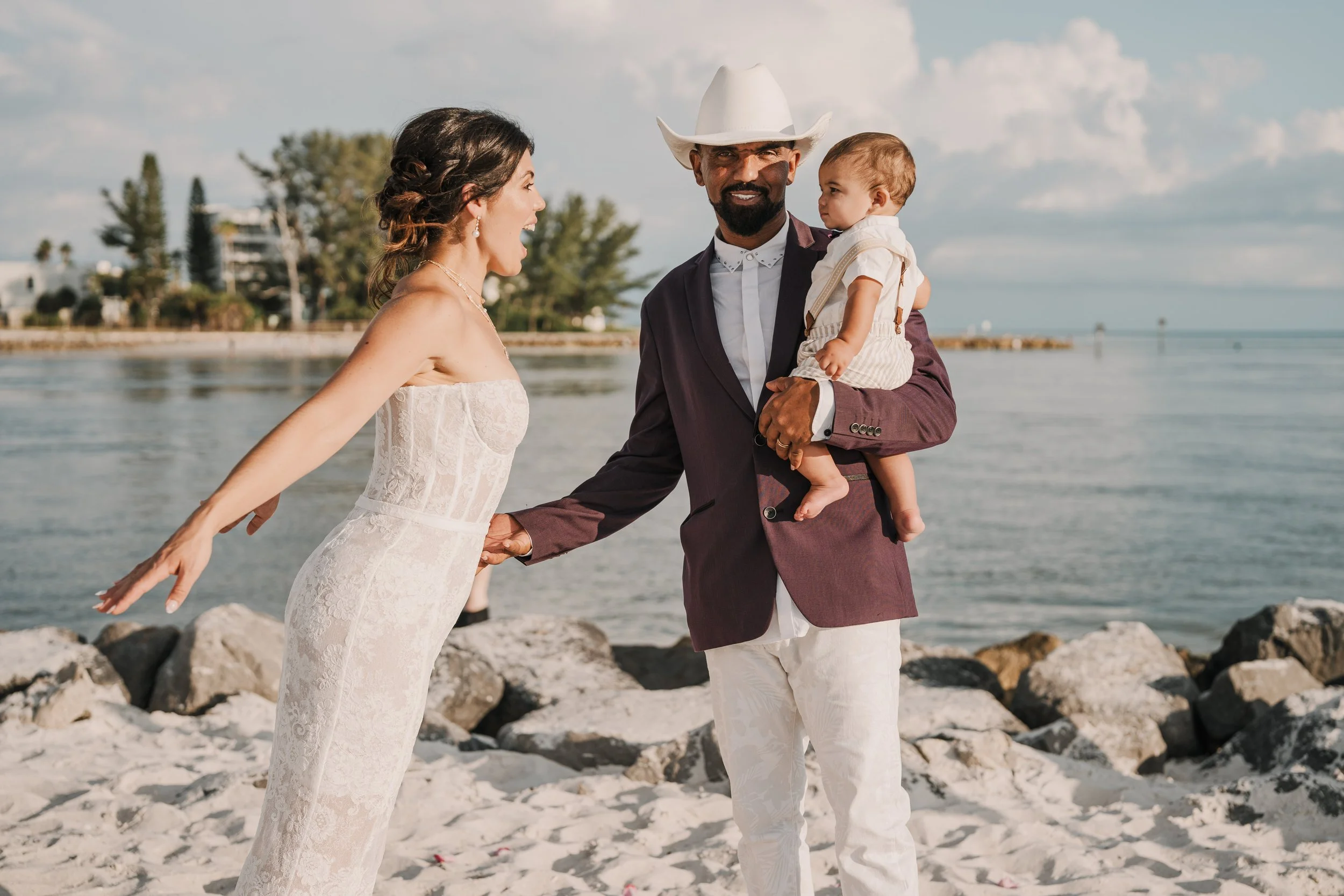 A bride and groom holding hands on the beach with a man holding a young child, sky and ocean in the background