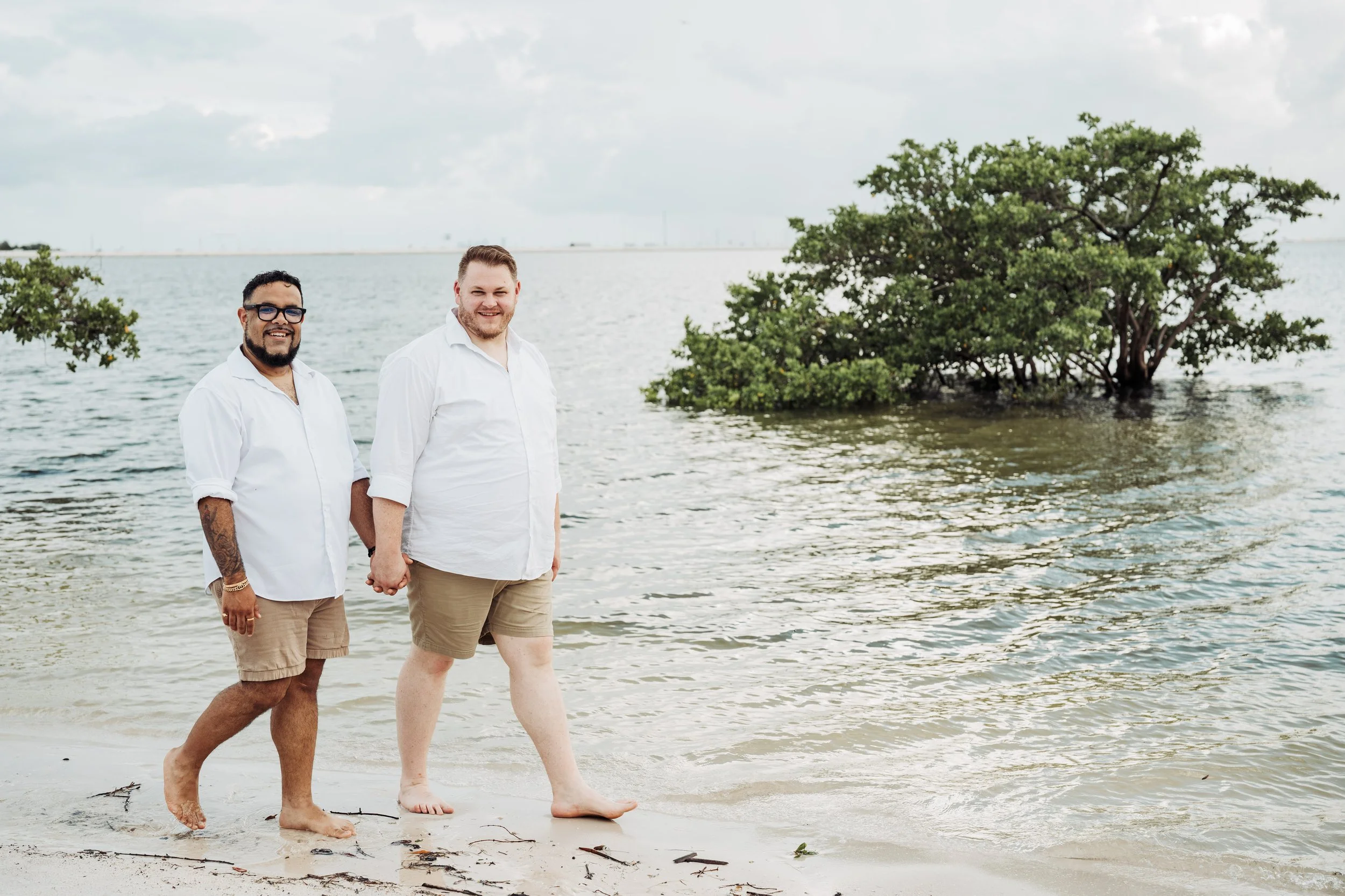 Two men holding hands and walking barefoot along the shoreline of a beach with water and trees in the background.