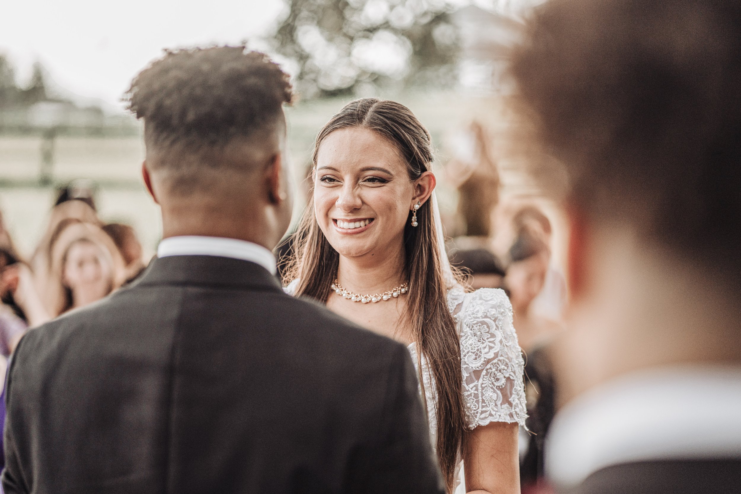 A woman with long brown hair smiling during a wedding ceremony, looking at a man with short dark hair, outdoors with blurred guests in the background.