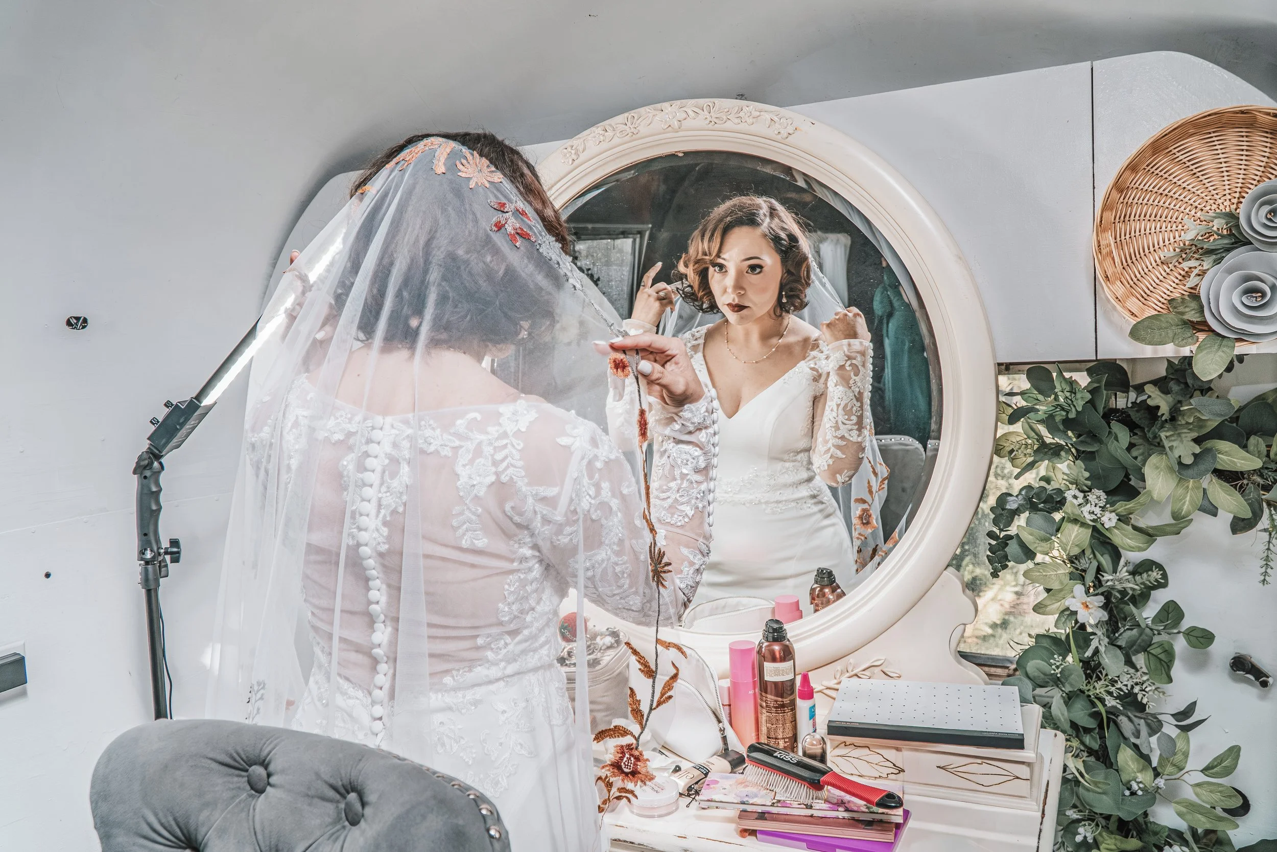 A woman in a white wedding dress with lace details is looking at herself in a mirror as she gets ready, with a makeup artist assisting her. The vanity area is decorated with green foliage, flowers, and various beauty products.