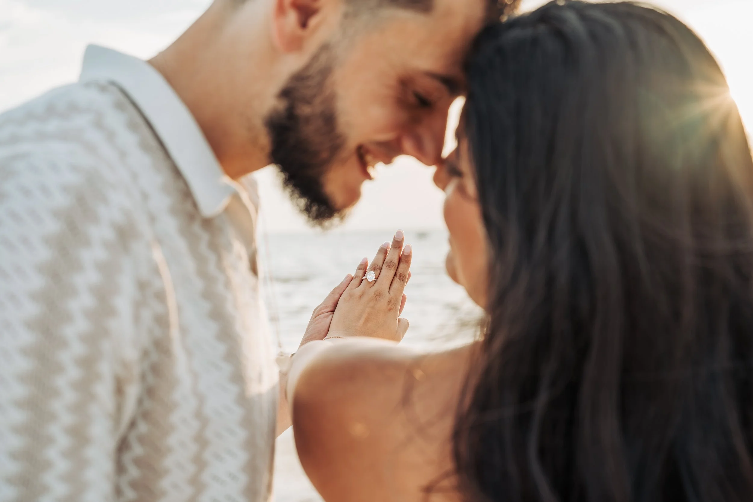A couple at the beach with their foreheads touching, the man wearing a white shirt and the woman with long dark hair, sun shining, and the woman showing a ring on her finger.