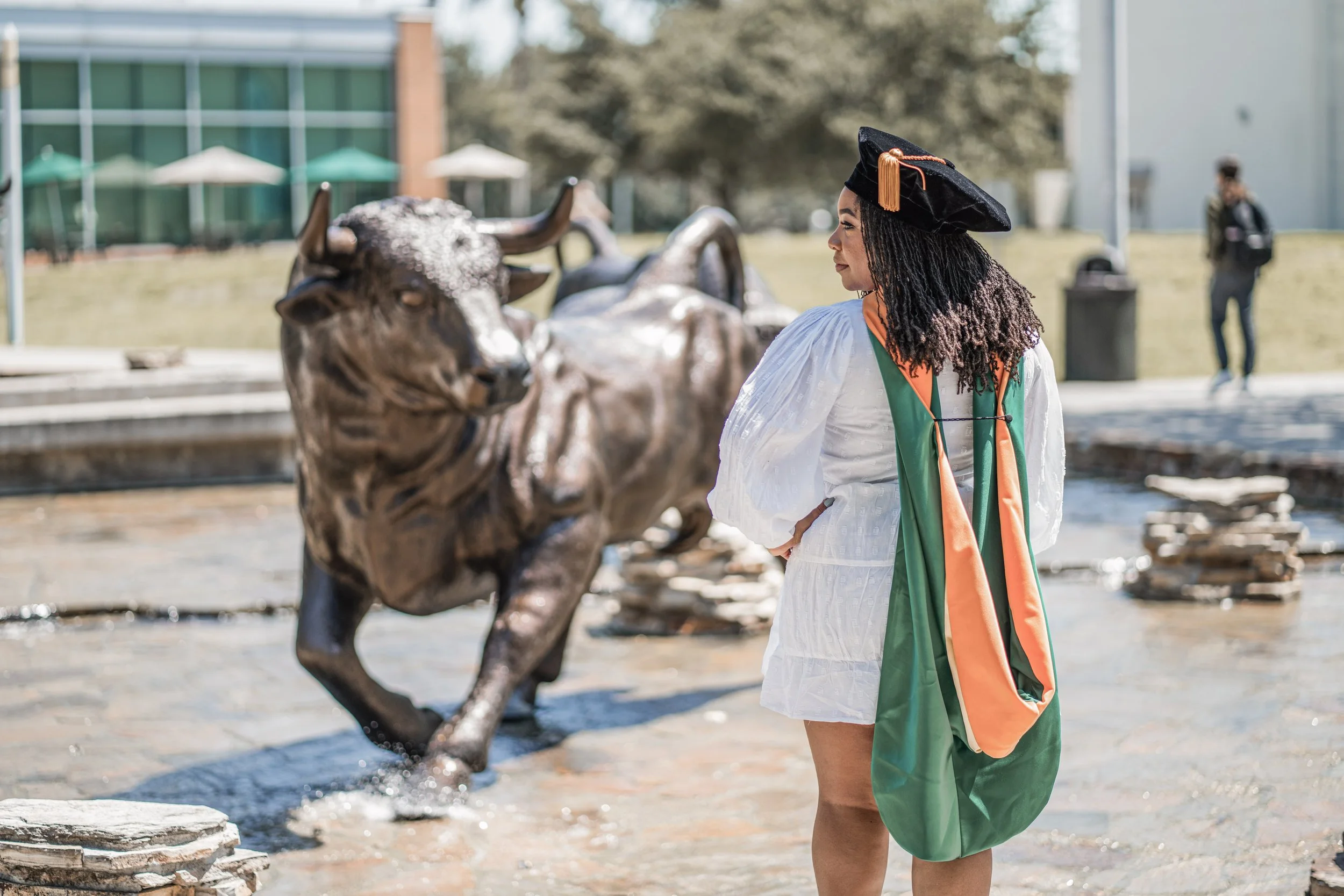 A woman in a graduation cap and gown standing outdoors near a fountain with a large bronze buffalo sculpture, with trees and modern buildings in the background.