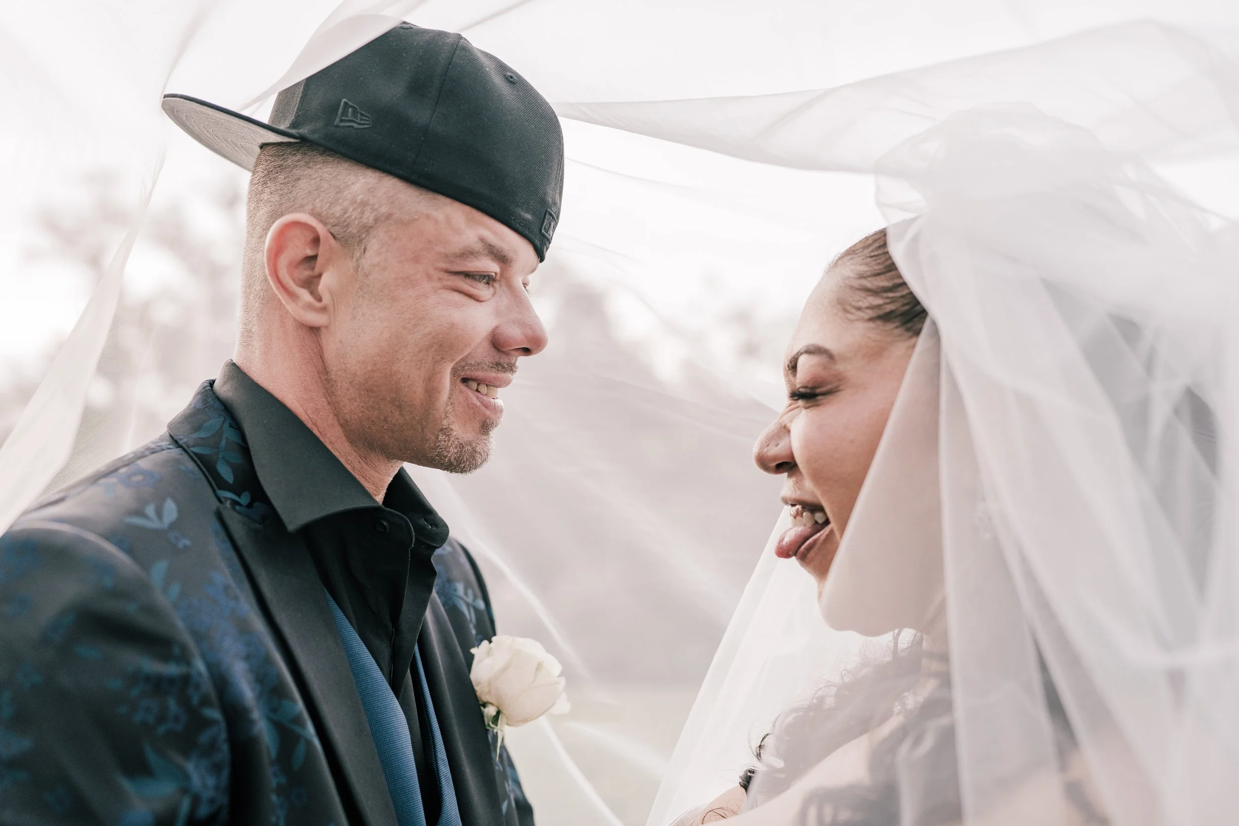 A smiling man and woman sharing a tender moment under white fabric, possibly during a wedding ceremony.