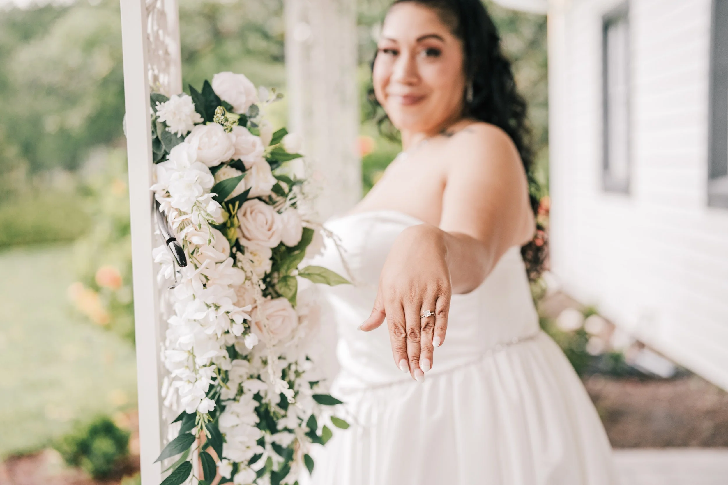 A bride showing her wedding ring while standing beside a floral arrangement of white and pale pink roses and greenery.
