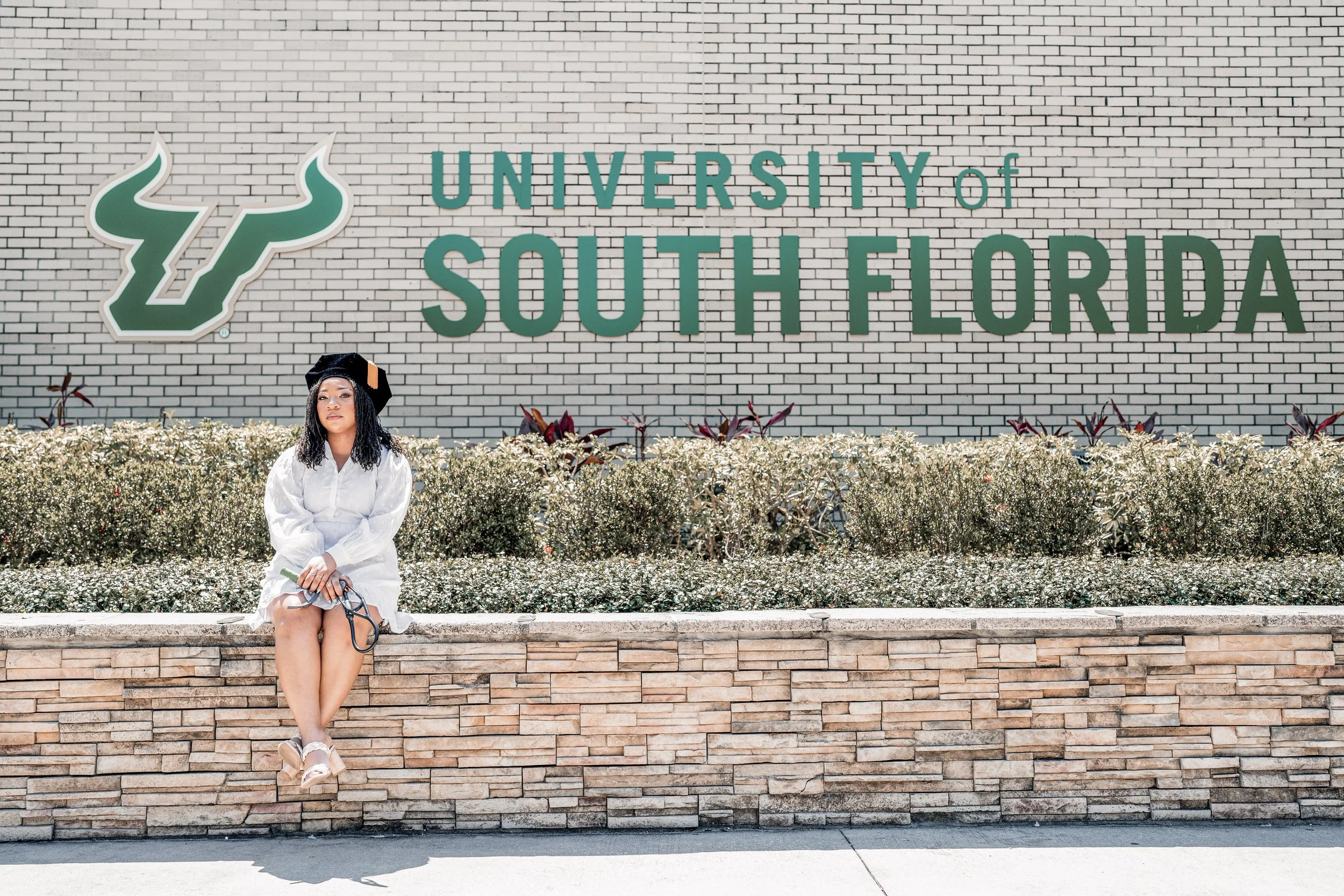 A woman in a white dress and a graduation cap sitting on a bench in front of a large sign that reads 'University of South Florida' with a logo on a brick wall background.