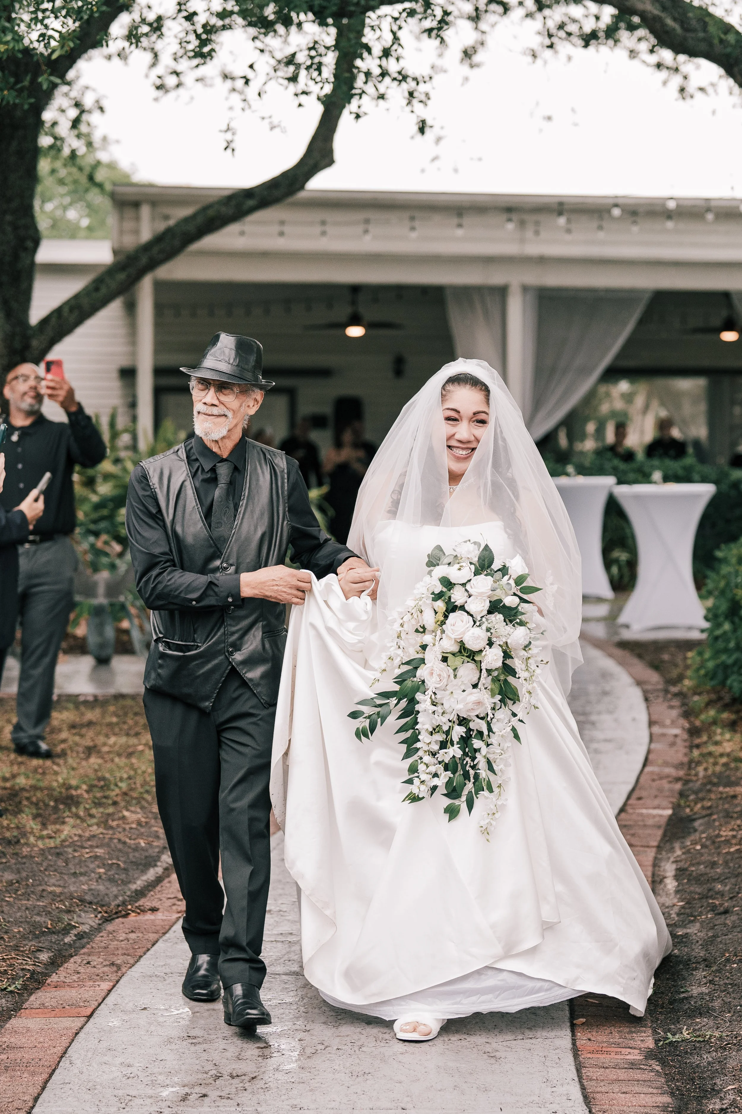 A bride in a white wedding dress and veil holding a bouquet, walking outside with a man in dark clothing and a fedora hat, while people in the background take photos at a wedding reception.