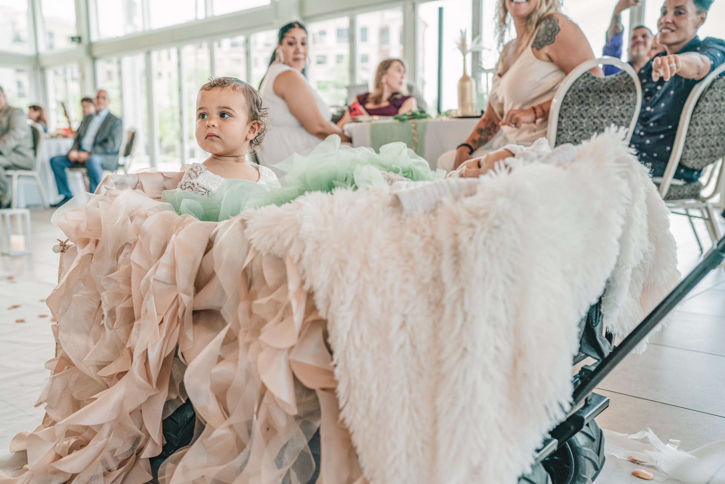 A young girl in a white dress with lace details sitting in a decorated stroller with pastel-colored tissue paper and furry fabric, at a social gathering with people seated at tables and a glass wall in the background.