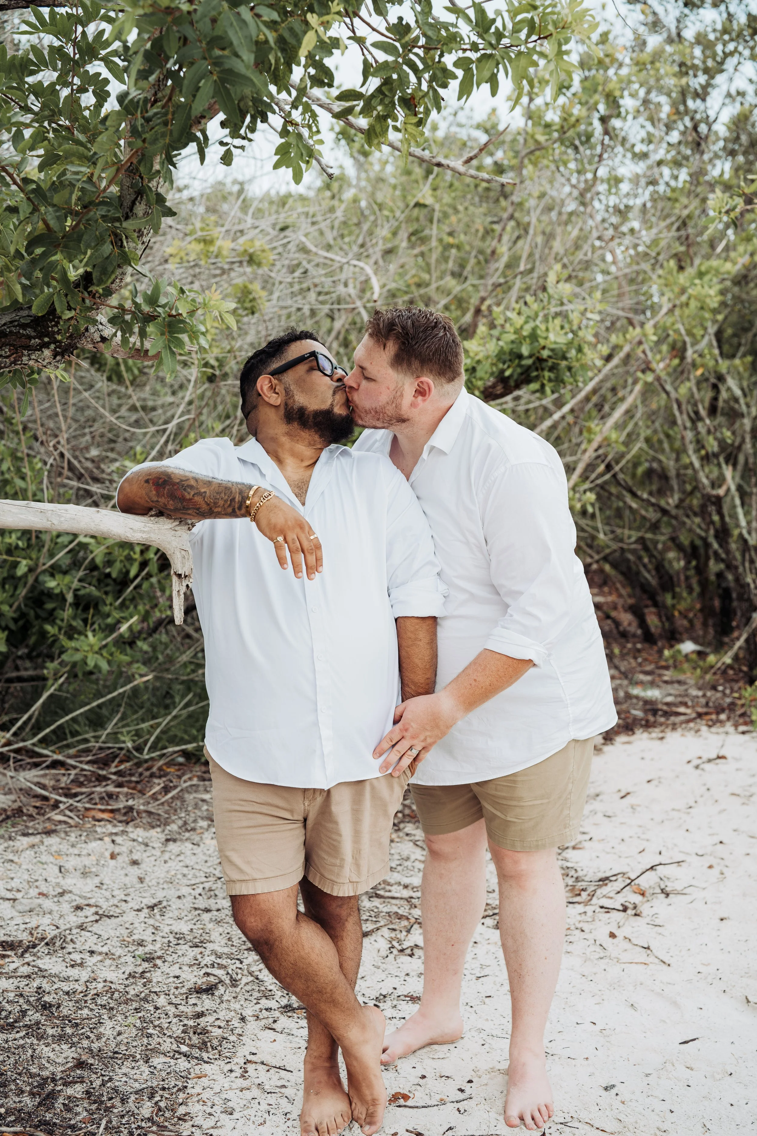 Two men sharing a kiss on a beach surrounded by trees. One man has dark hair, glasses, and tattoos, and the other has light hair. Both are dressed in white shirts and beige shorts.