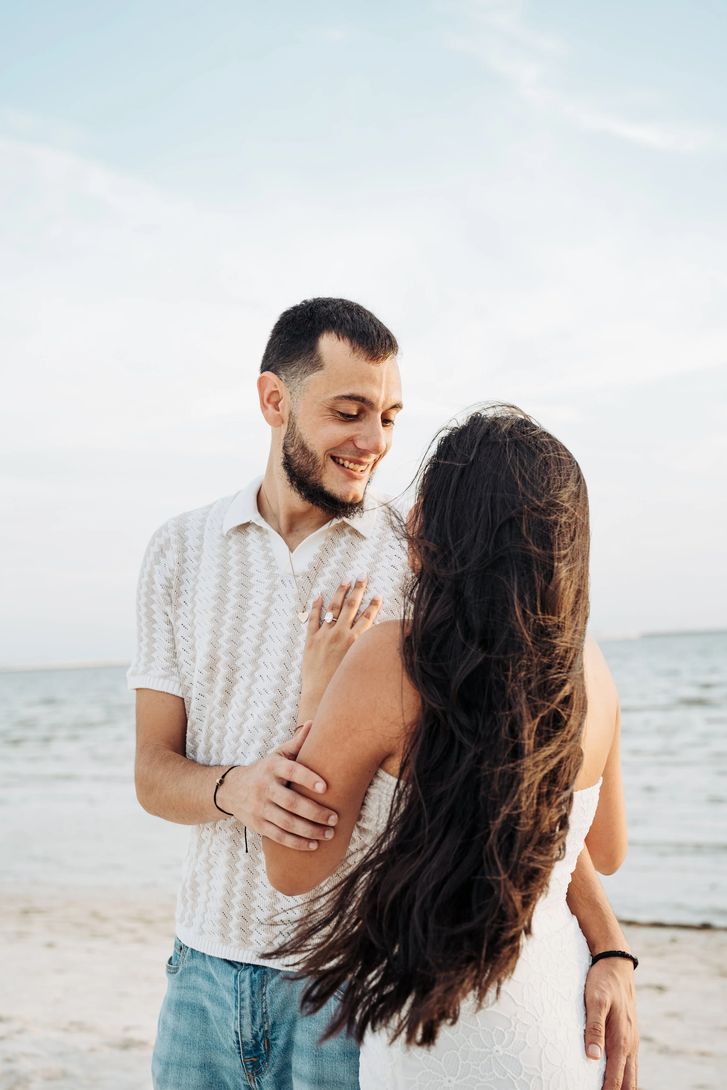A smiling man and woman standing on a beach, embracing each other with a body of water and cloudy sky in the background.
