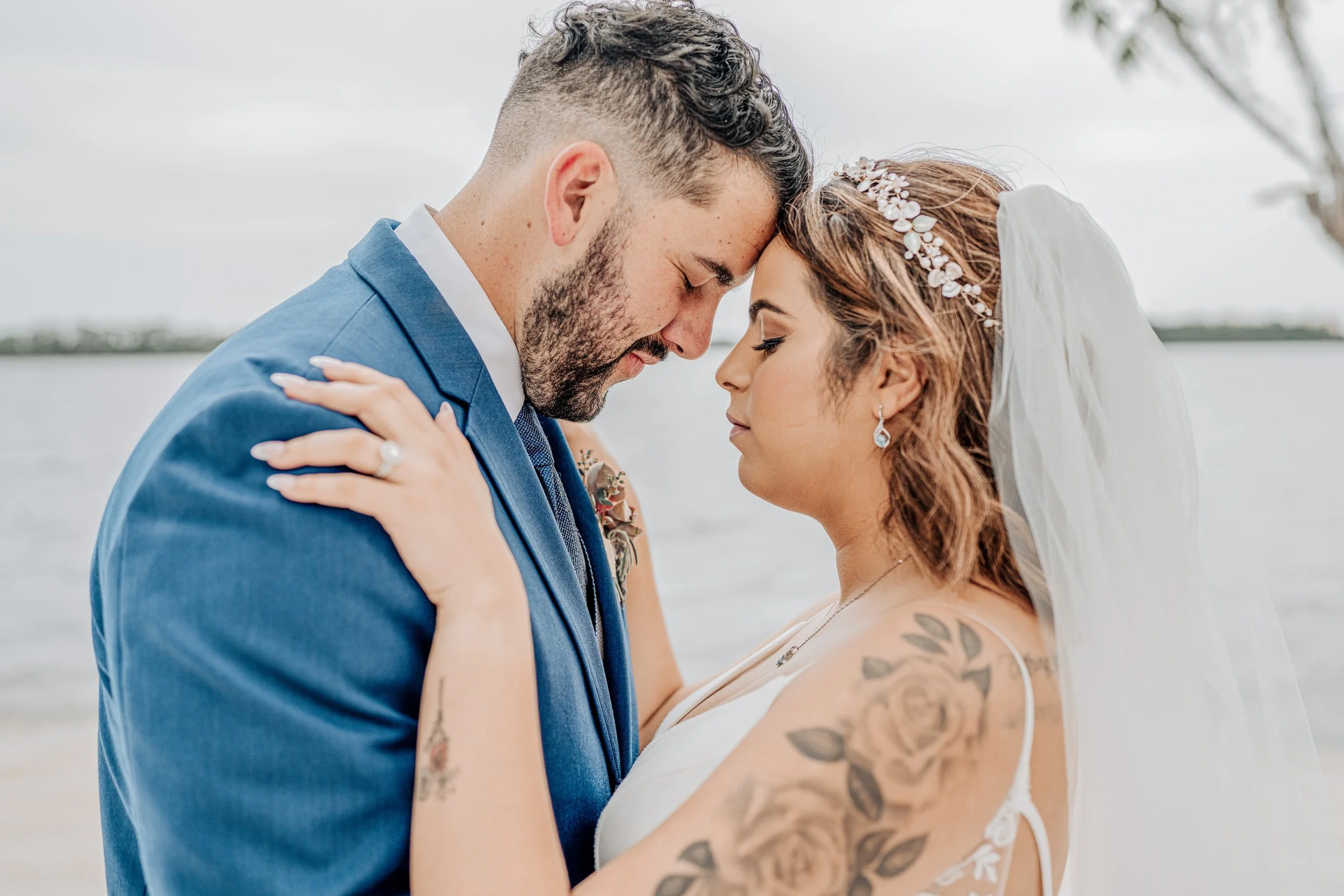 A couple, dressed in wedding attire, with foreheads touching and eyes closed, embracing during a wedding ceremony near a lake or river under an overcast sky.