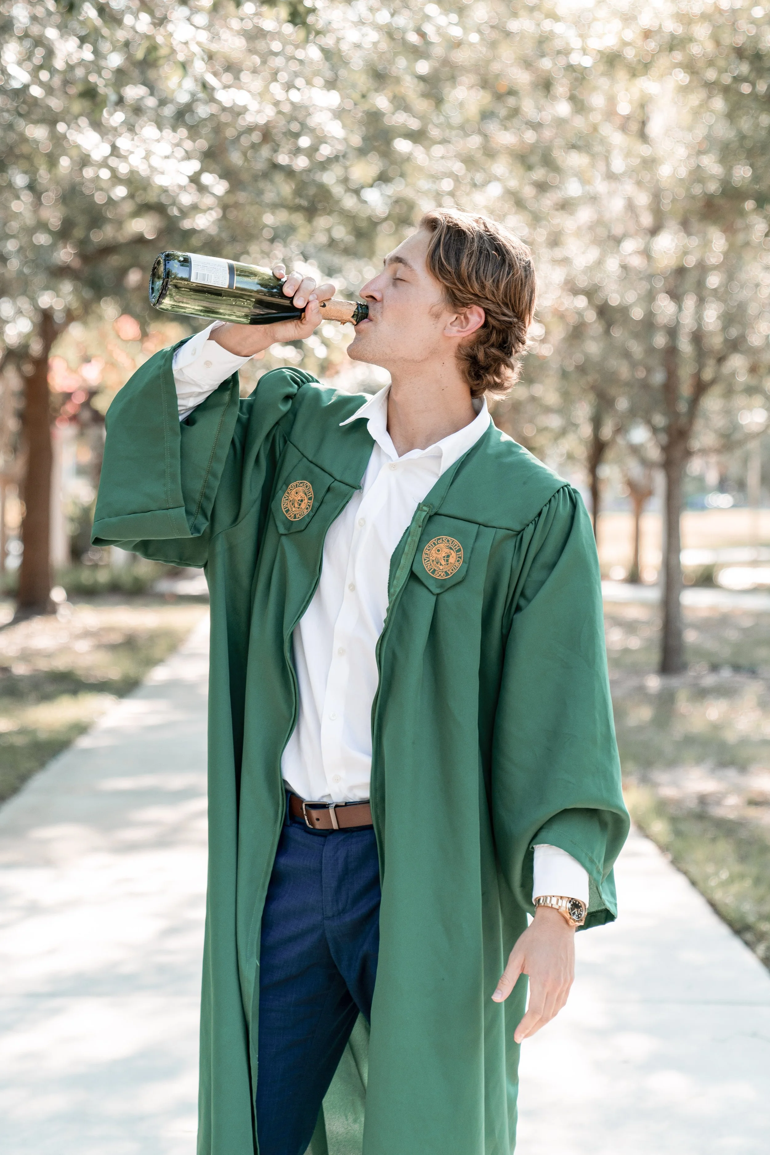 A young man in a green graduation gown and white shirt drinking from a bottle outdoors on a sunny day.