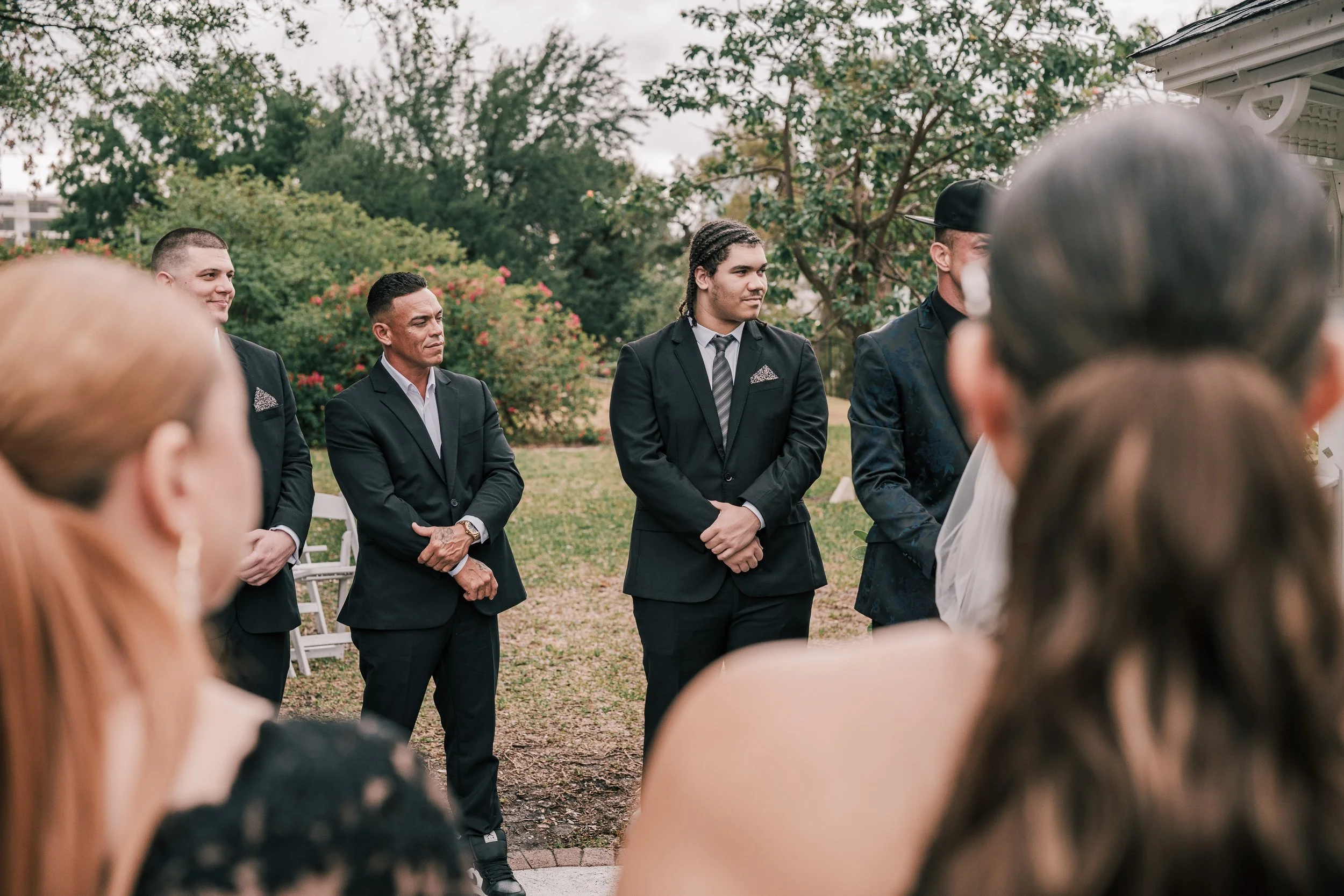 A group of men dressed in formal suits, standing outdoors at a gathering, with trees and bushes in the background.